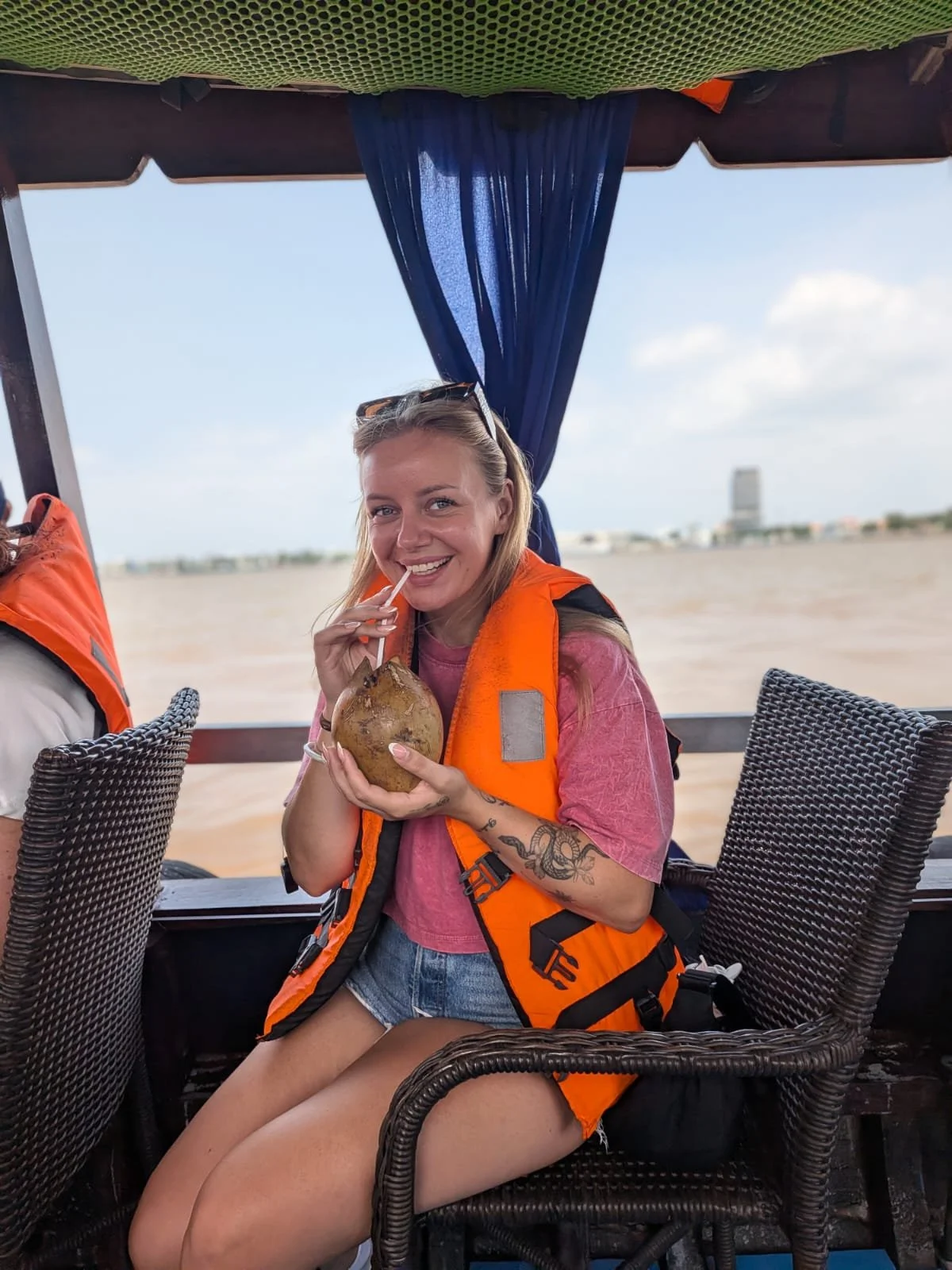Junge Frau mit Orange Rettungsweste sitzt auf einem Boot und trinkt aus einem Strohhalm, hält eine Kokosnuss in der Hand, im Hintergrund Wasser und eine Stadt am Horizont.