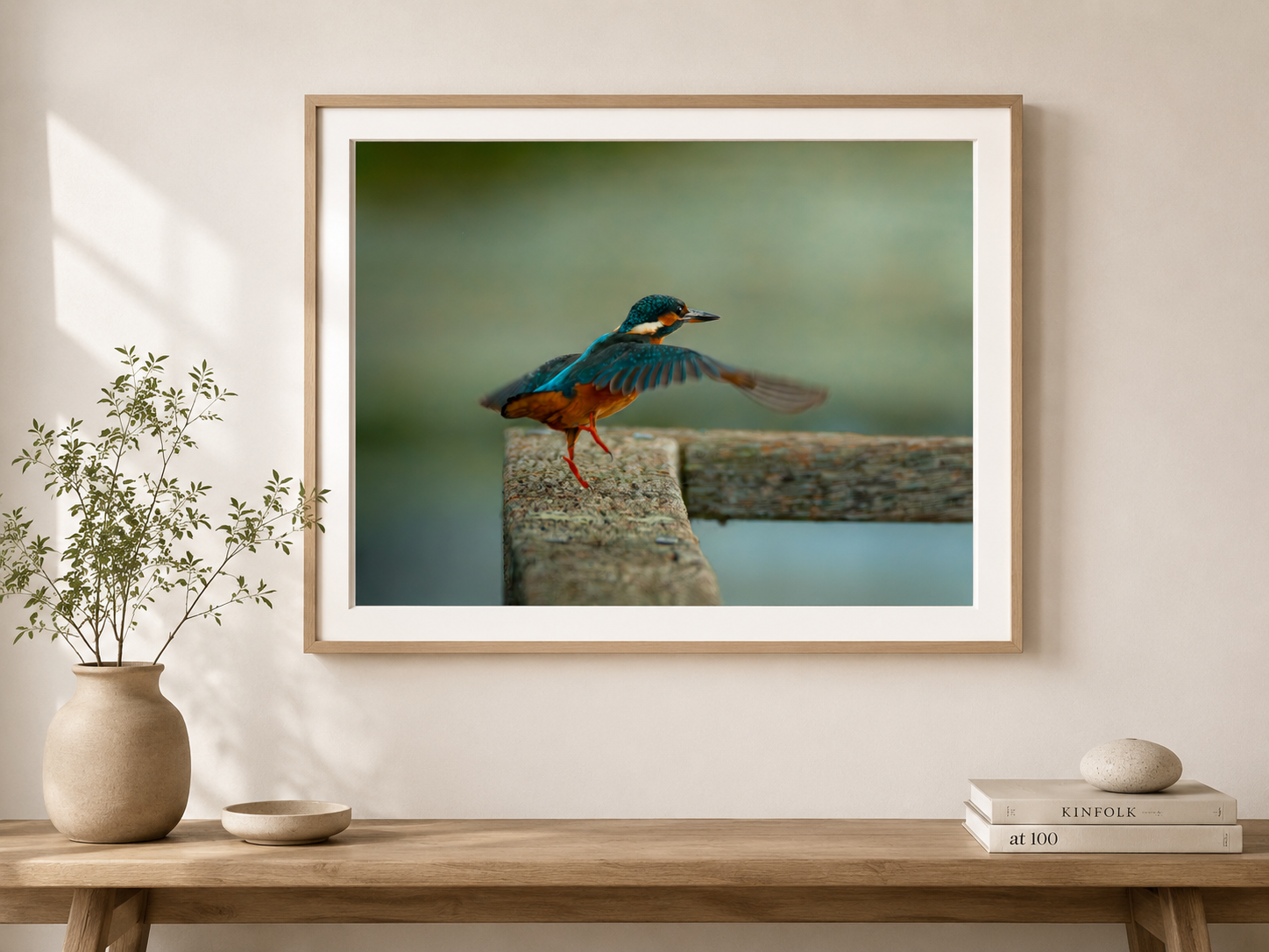 A framed photograph of a bird, possibly a kingfisher, perched on a wooden dock with one foot raised, with a blurred green background. The frame is on a beige wall, above a wooden table with a potted plant, a small dish, and two stacked books.