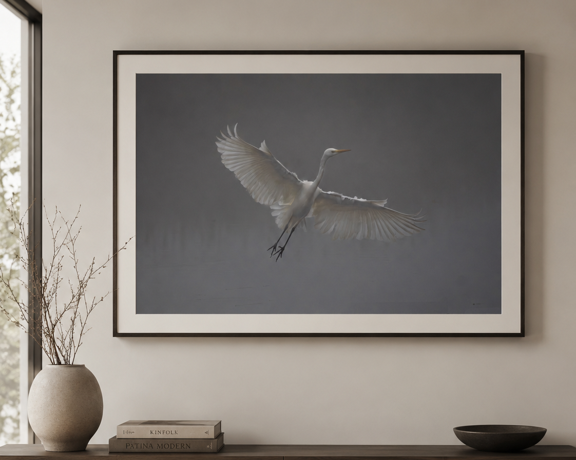 A photograph of a white heron flying in front of a dark grey sky, framed and hung on a wall above a wooden side table with decorative items.