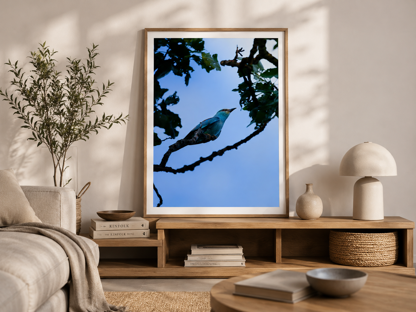 Decorative living room with framed bird art, beige sofa with throw blanket, green houseplant, wooden side tables with books and vases, and a table lamp, sunlit with neutral tones.