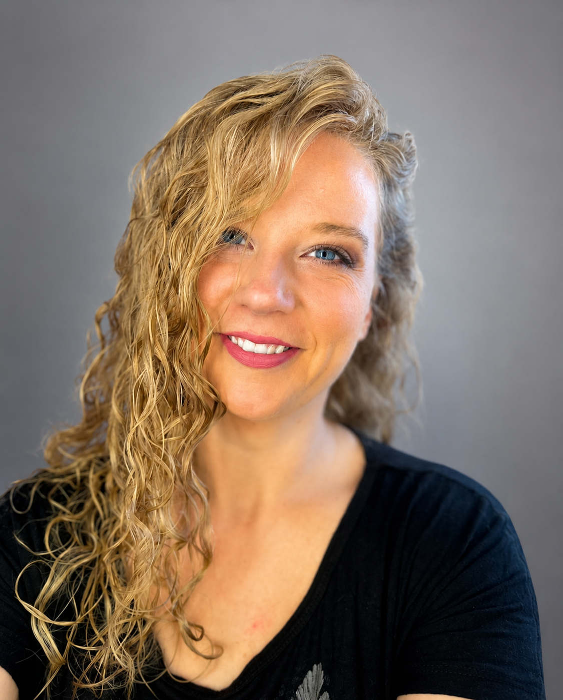 A woman with blonde, curly hair, blue eyes, and a black shirt, smiling against a gray background.