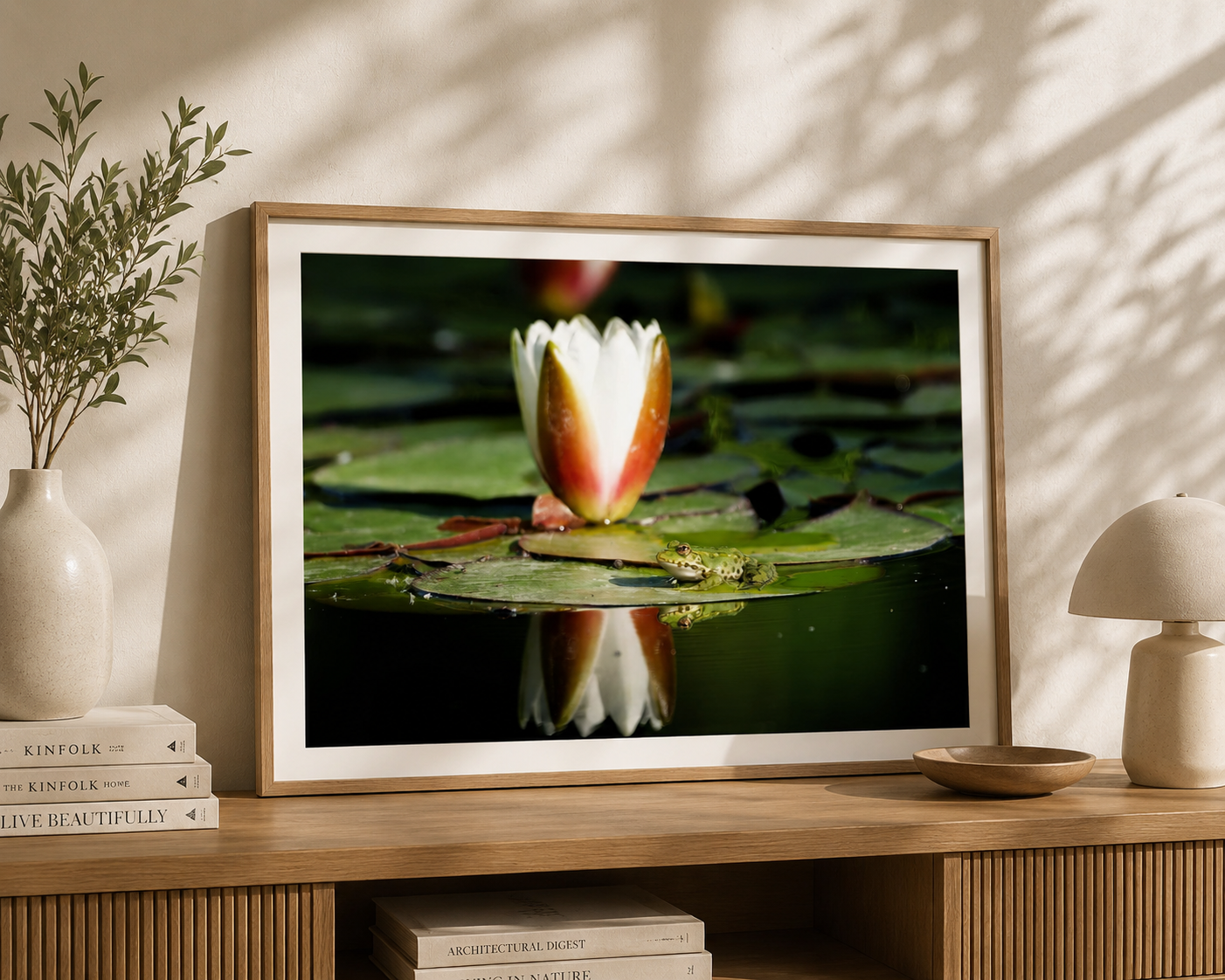 Framed photograph of a white water lily and a small frog on a lily pad, sitting on a wooden surface with decorative vases, a lamp, and books nearby.