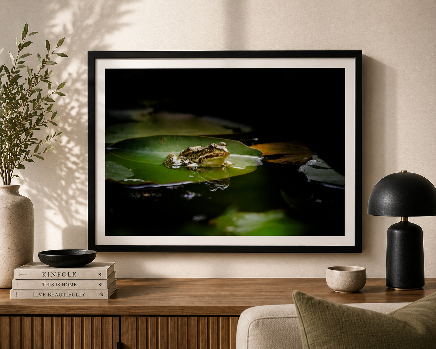 Framed photograph of a frog sitting on a green lily pad in water, hung on a beige wall in a living room. The room has a wooden sideboard with stacked books, a black ceramic bowl, a beige vase with green foliage, a small beige bowl, and a black and go