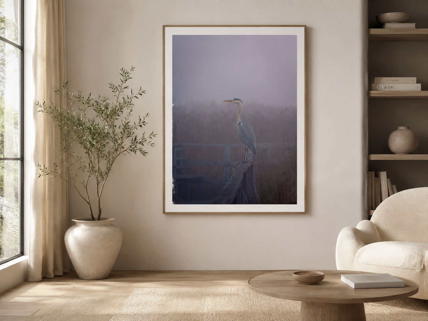 A living room with a large framed photograph of a heron on a dock, an off-white armchair, a small wooden coffee table with a bowl and a book, a tall potted plant in a white ceramic pot, beige curtains, and built-in shelves with decorative objects.