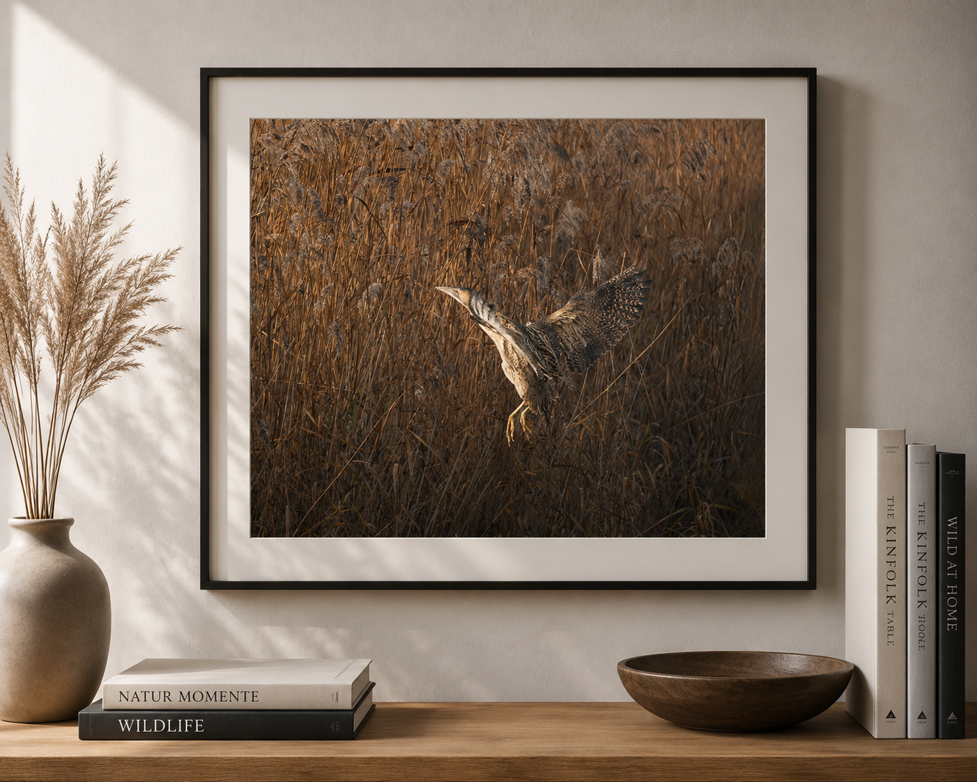 A framed photograph of a heron flying over tall brown grasses, hanging on a wall in a room with books and decorative items on a wooden shelf.