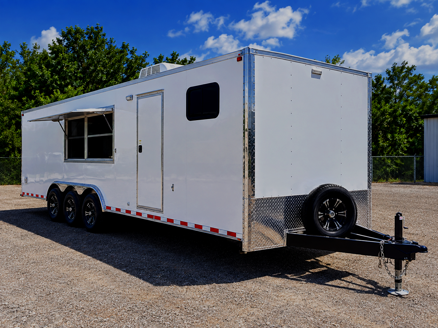 White enclosed trailer with black wheels, a side window, and a side door, parked on gravel with a blue sky and trees in the background.