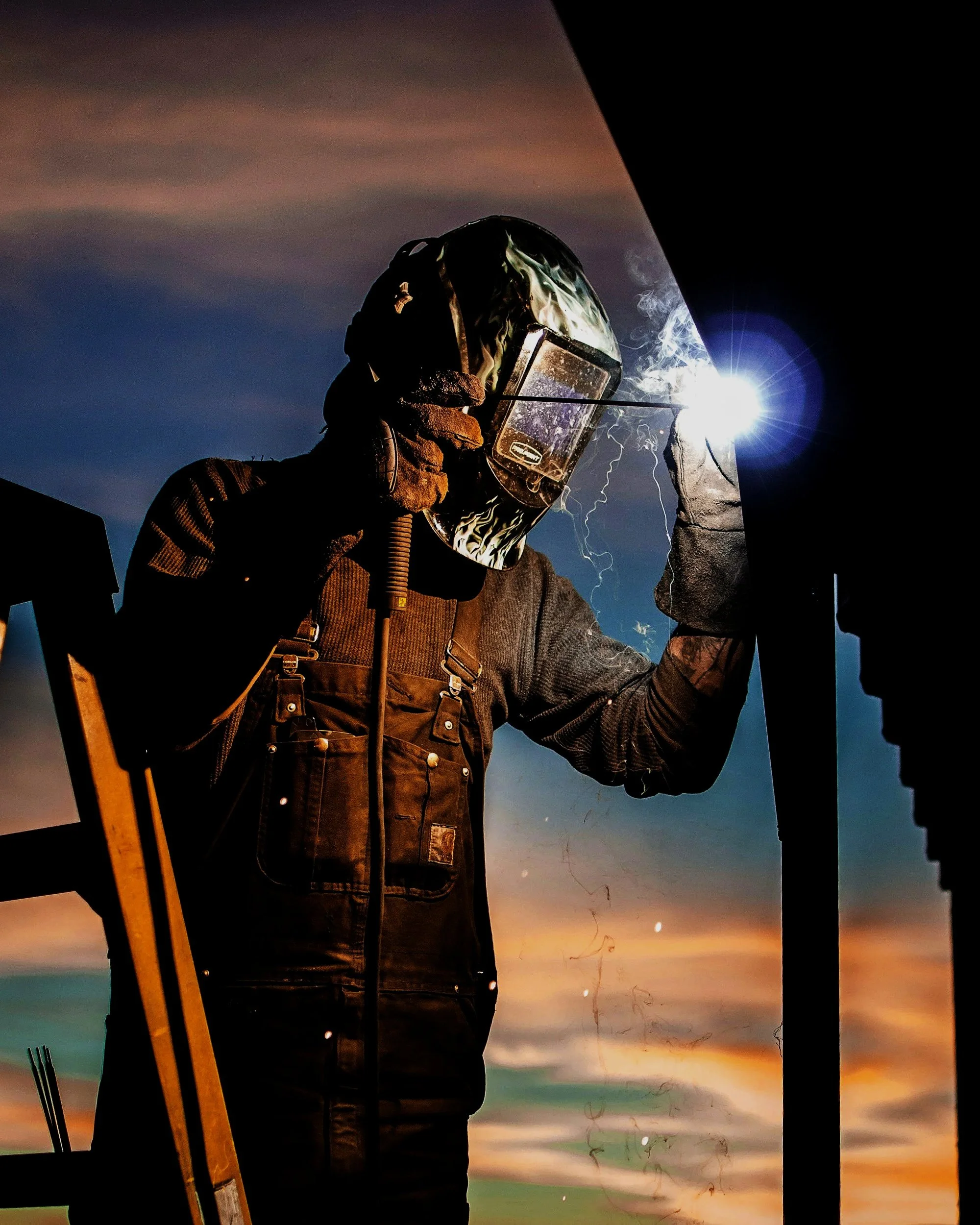 A person welding outdoors at sunset, wearing protective gear including a welding helmet, gloves, and a jacket, with sparks and smoke visible from the welding process.
