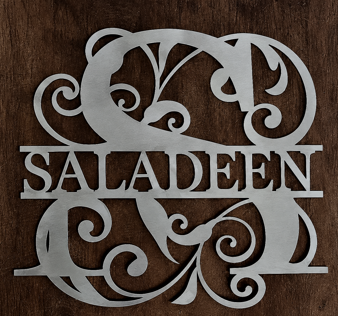 Metal wall decor with the word 'Saladen' in capital letters, surrounded by ornate swirling floral patterns, on a wooden background.