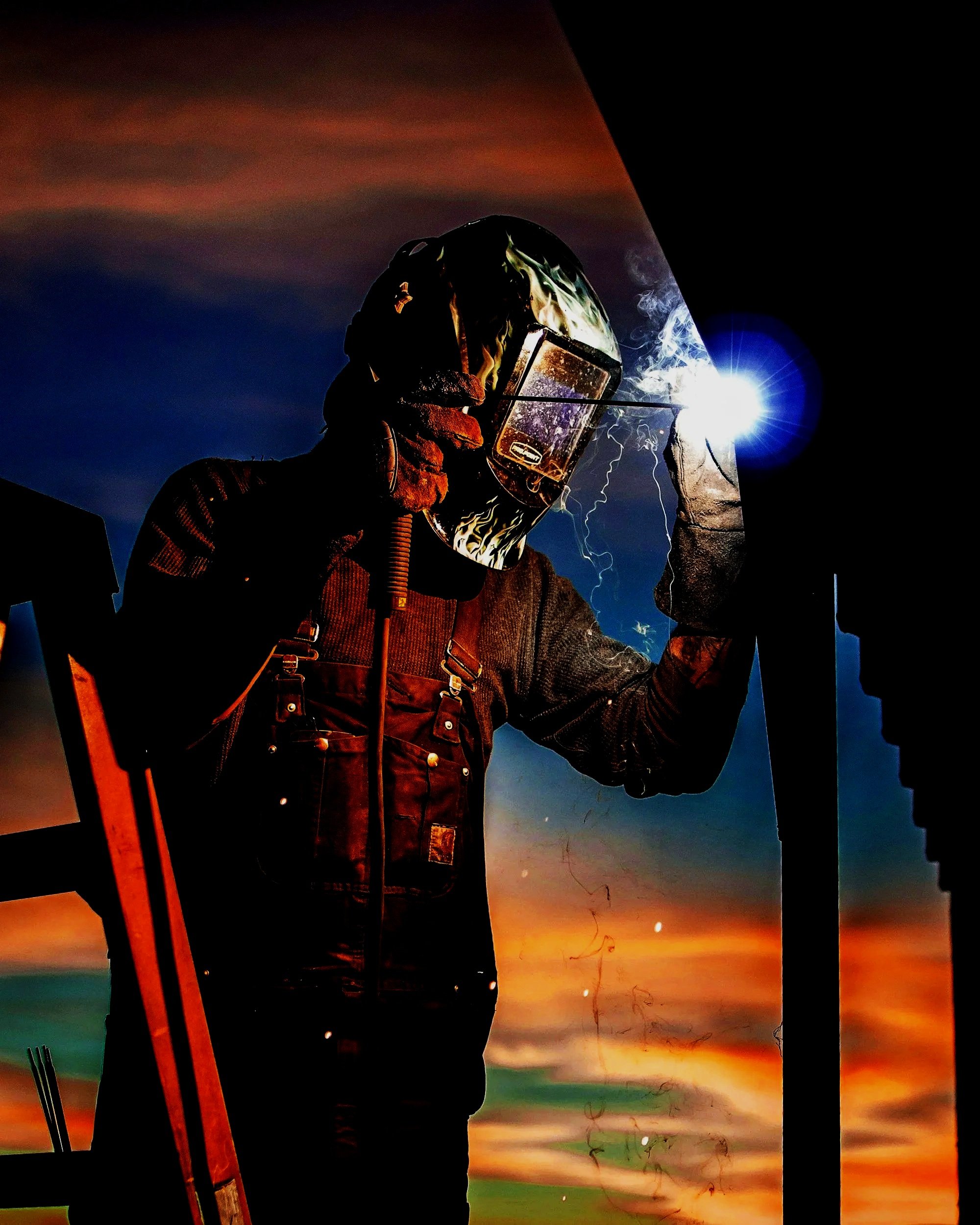 Silhouette of a welder wearing a protective helmet and gloves, welding outdoors at sunset with sparks and smoke, against colorful sky.
