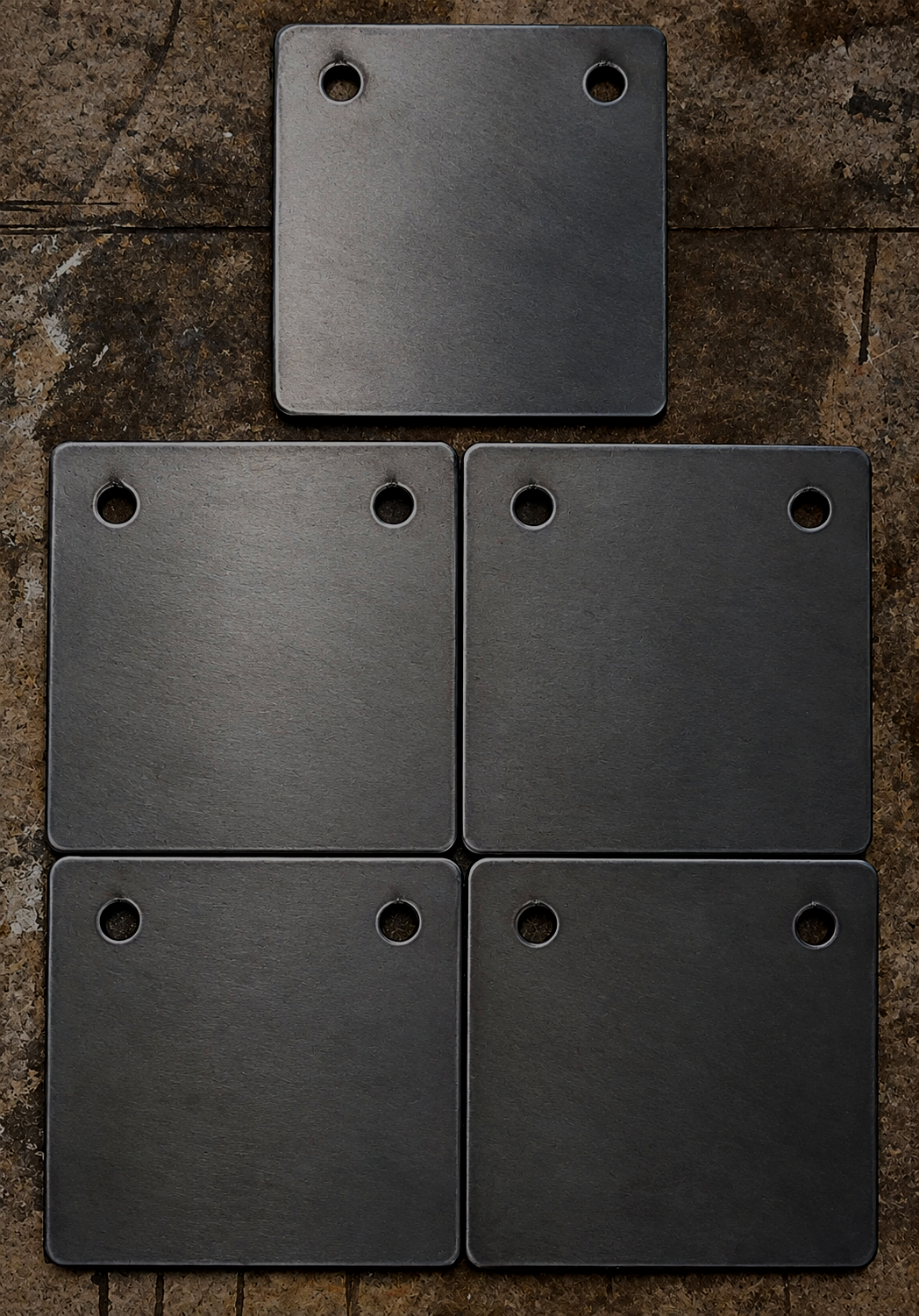 Five black square metal plates with rounded corners and four small circular holes, arranged on a rusty brown surface, with one plate positioned at the top and four below it.