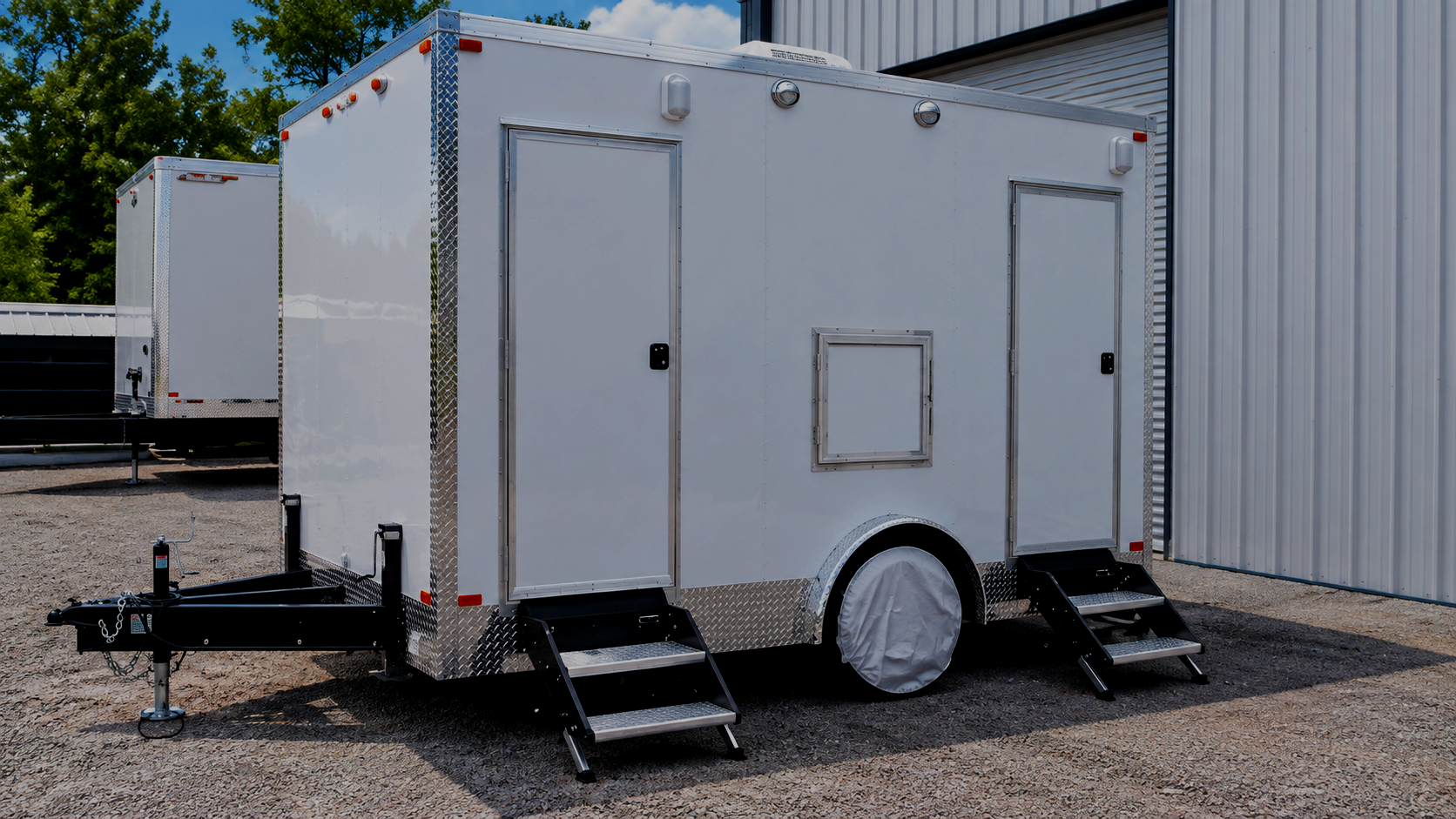 A white mobile food or utility trailer with steps on both sides, parked outdoors near a building with a metal exterior wall, surrounded by gravel and trees in the background.