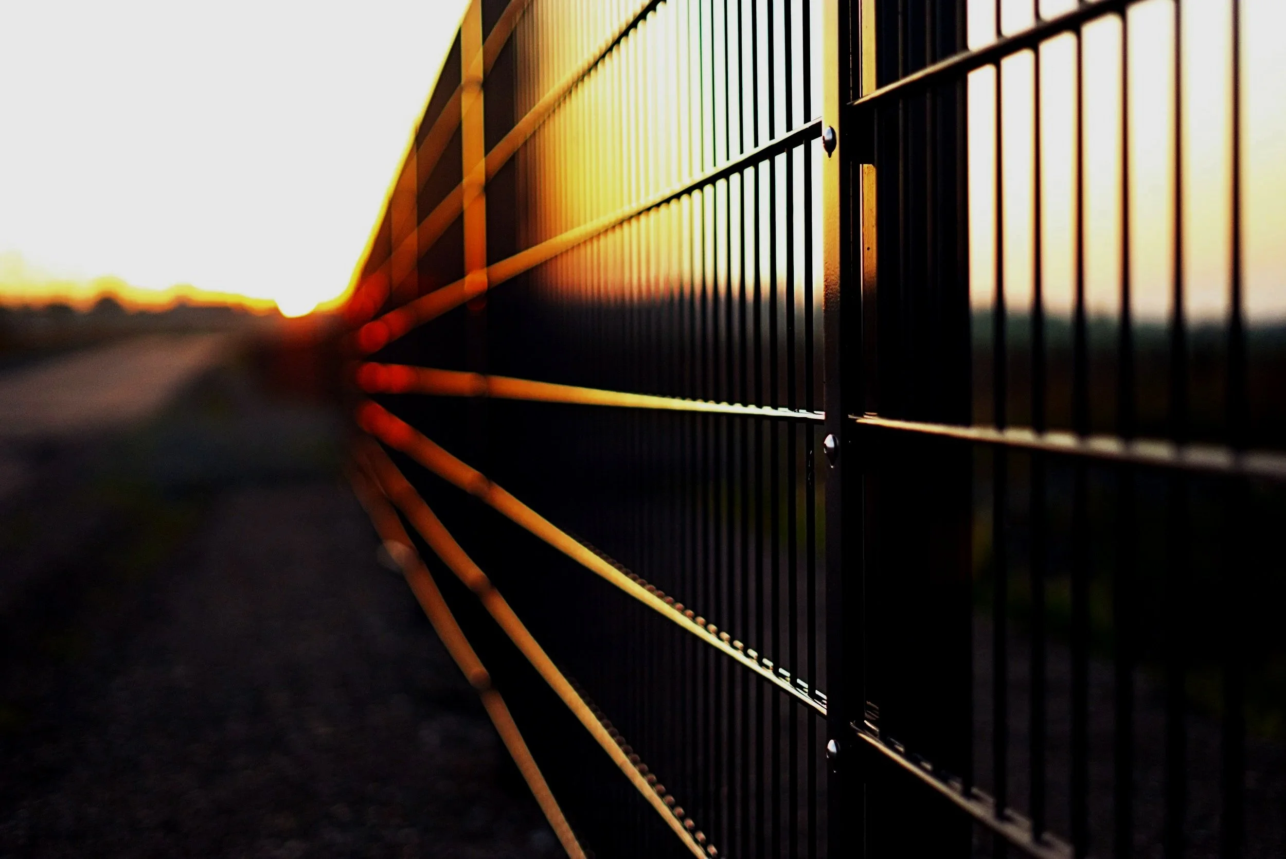 Close-up of a black metal fence with yellow horizontal bars during sunset, creating a colorful contrast against the bright sky in the background.