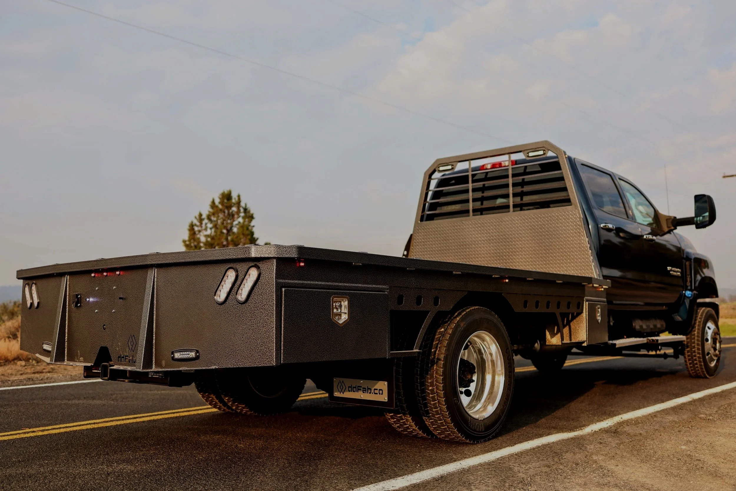 Black flatbed utility truck with a diamond plated metal cab guard, parked on a road with yellow lane markings, under a partly cloudy sky, and trees in the background.