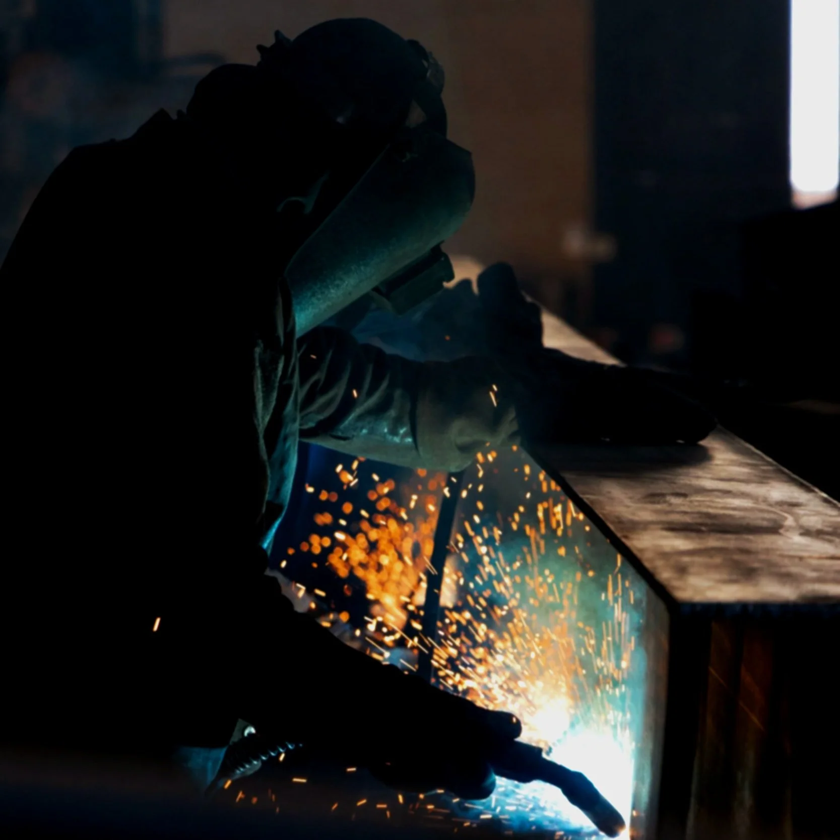 A person welding metal at a workbench, sparks flying from the welding process in a dimly lit workshop.