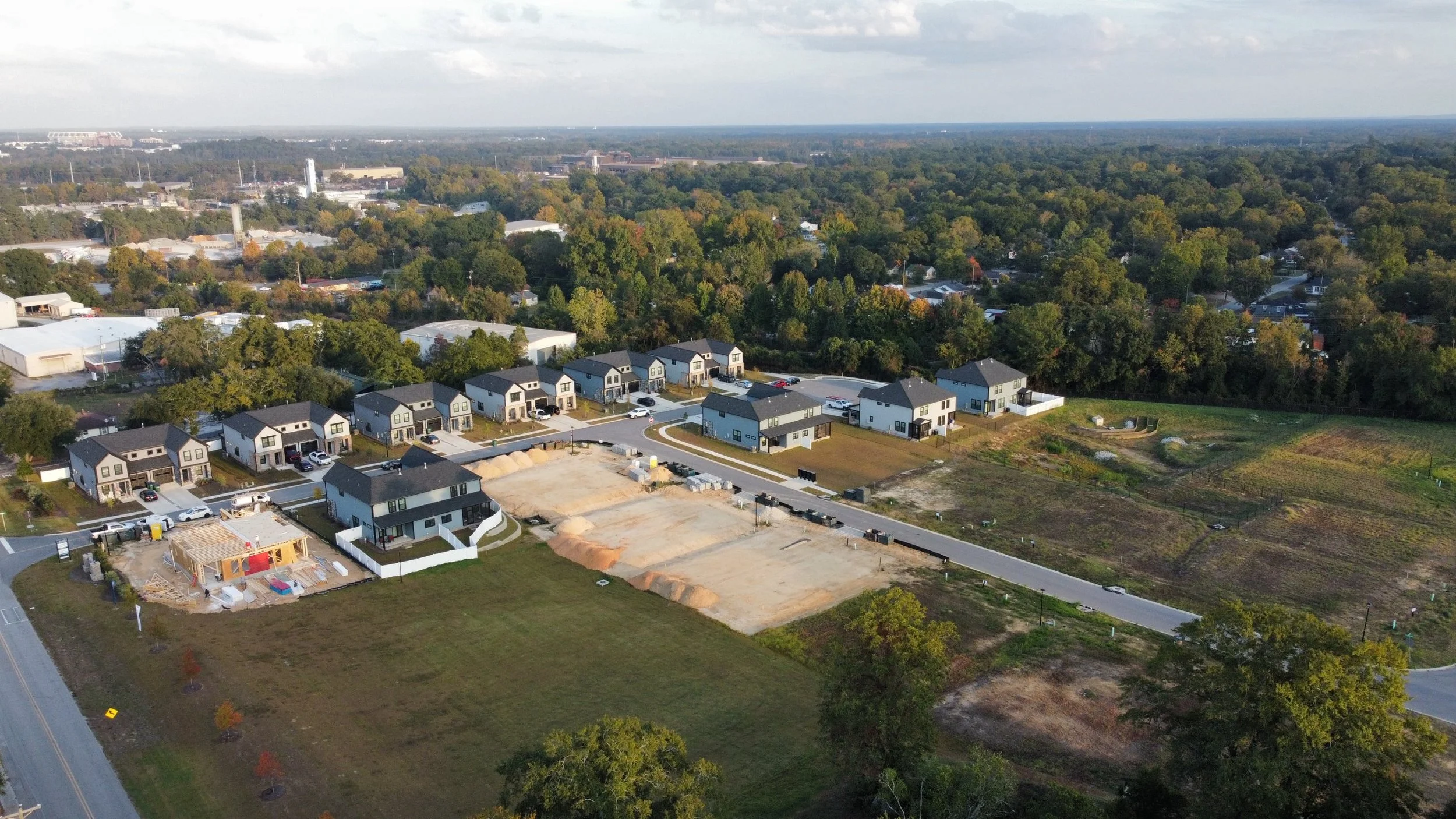 Aerial photo of a neighborhood under construction: finished homes along new streets, a house being framed, graded lots with sand piles, and a fenced stormwater basin.