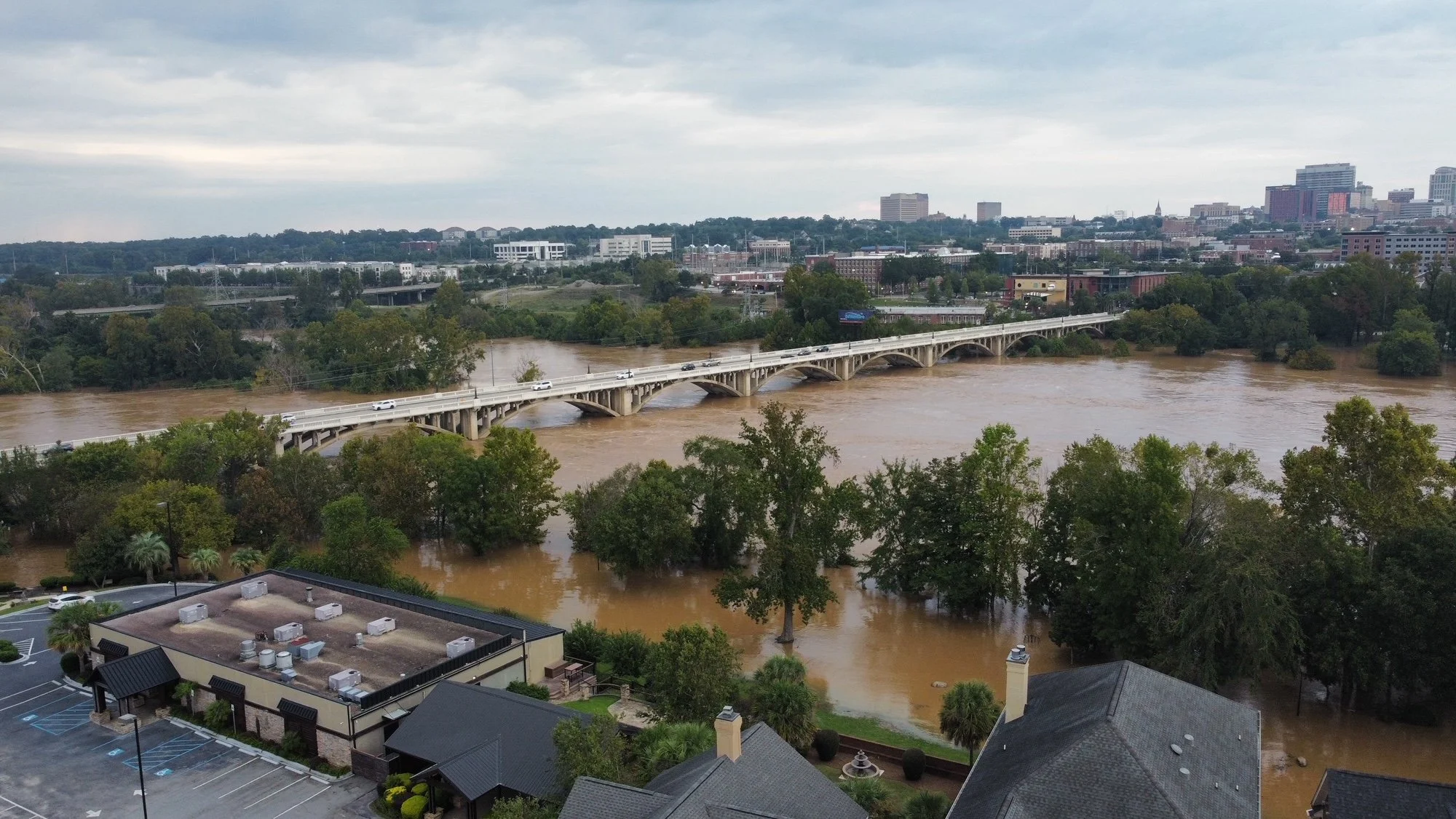 Drone view of the concrete arches of Gervais Street Bridge crossing the swollen Congaree River during Hurricane Helene flooding, with flooded riverfront trees and downtown Columbia in the background.