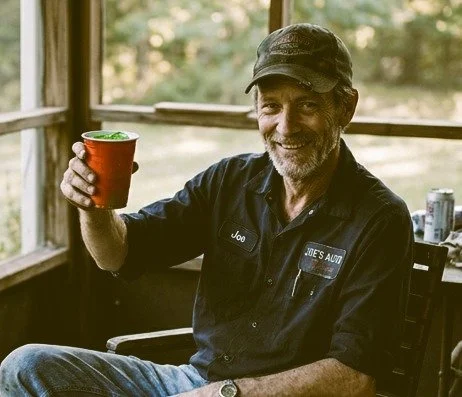 A man with a gray beard, wearing a black work shirt and cap, smiling and holding a red cup in a sunlit room.