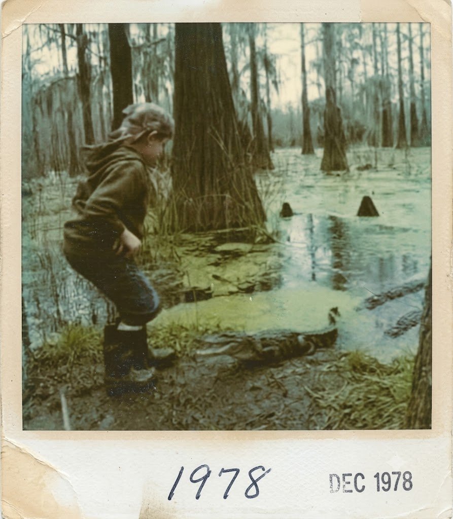A young girl standing in a muddy area near a swamp or wet forest, wearing a hoodie and rubber boots, with a snake on the ground in front of her. The background features tall trees and water, with some tree stumps rising above the water. The photo is dated December 1978.
