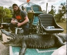 A man standing next to a large crocodile with a saddle and equipment on it, outdoors with trees and blue sky.