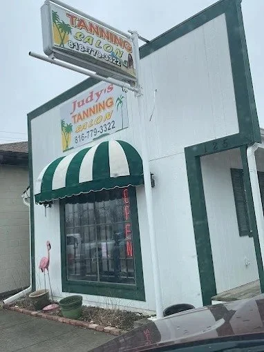 Street view of a tanning salon called 'Judy's Tanning' with a green and white striped awning over the window, a neon 'OPEN' sign in the window, and a sign on top of the building advertising tanning services and a phone number.