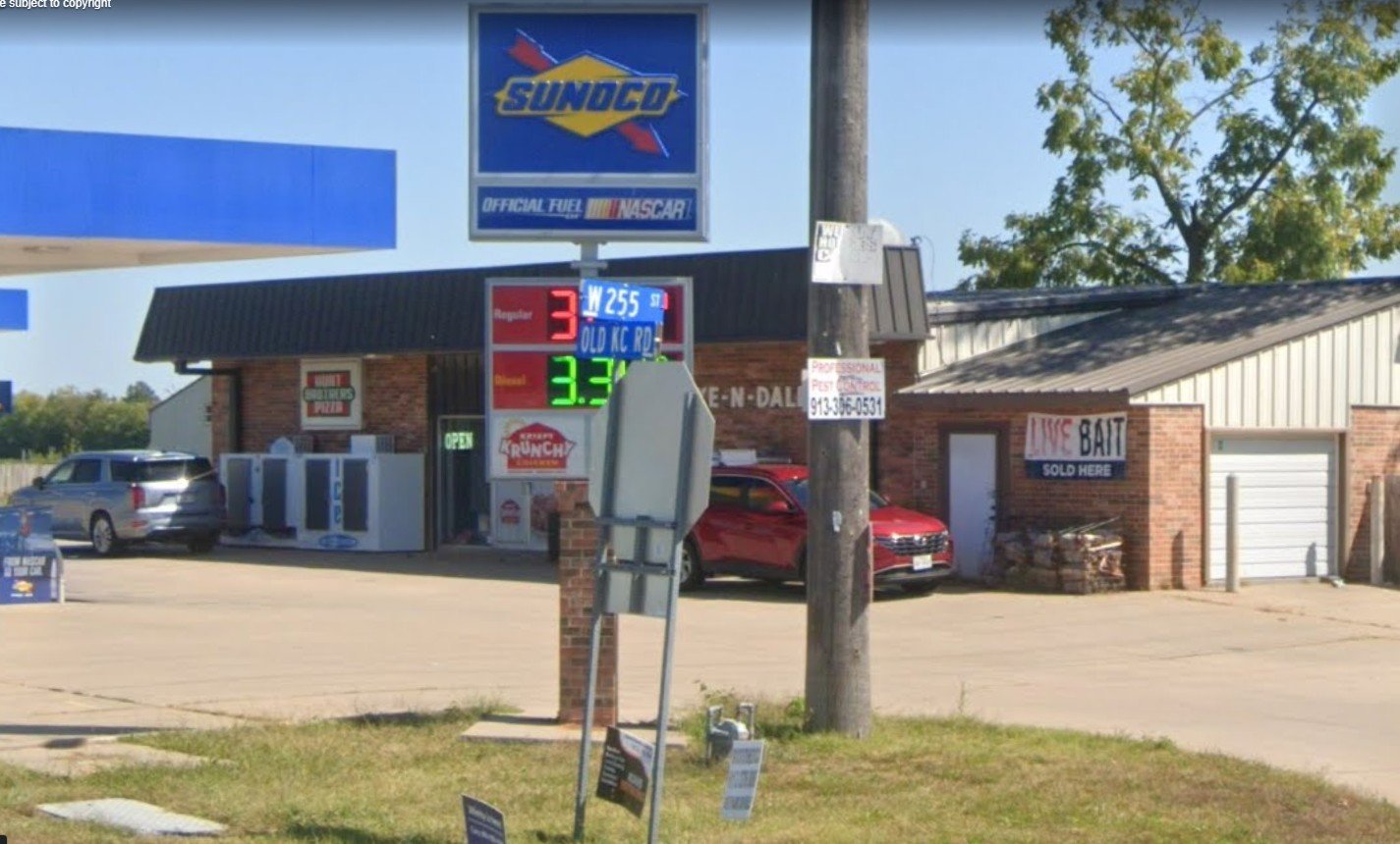 A Sunoco gas station with a digital sign displaying fuel prices, a red SUV parked near the store, and a sign advertising live bait sold on site. There are trees and a blue sky in the background.