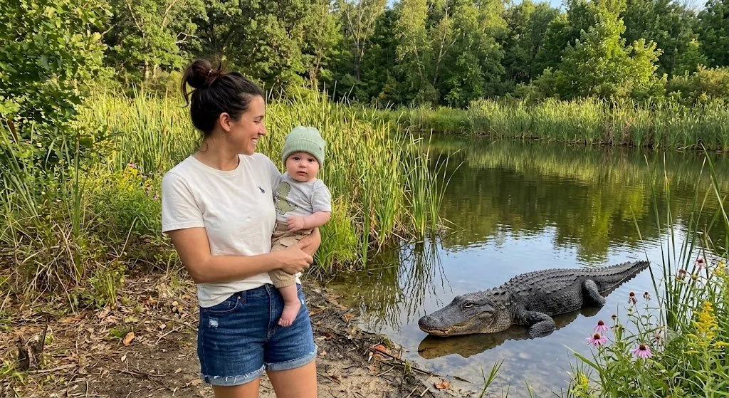 A woman holding a young child standing near a riverbank with an alligator in the water nearby, surrounded by greenery.