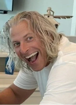 A man with long, curly blonde hair smiling excitedly in a room, possibly a hospital or clinic setting.