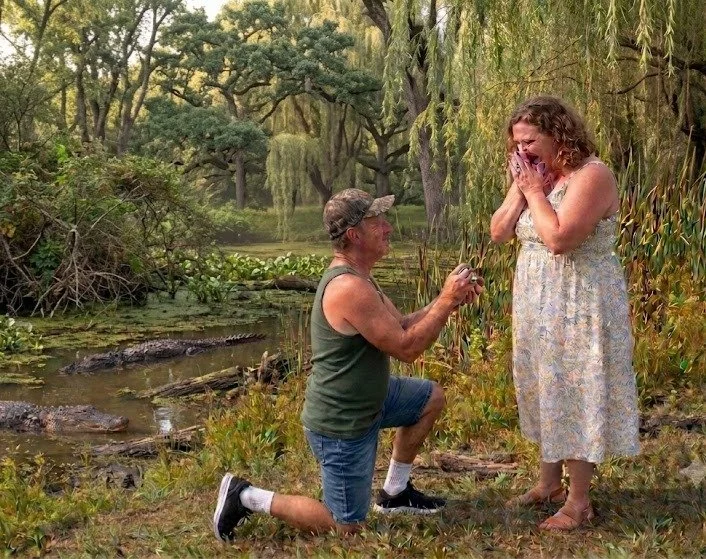 A man proposes to a woman outdoors near a pond surrounded by trees.