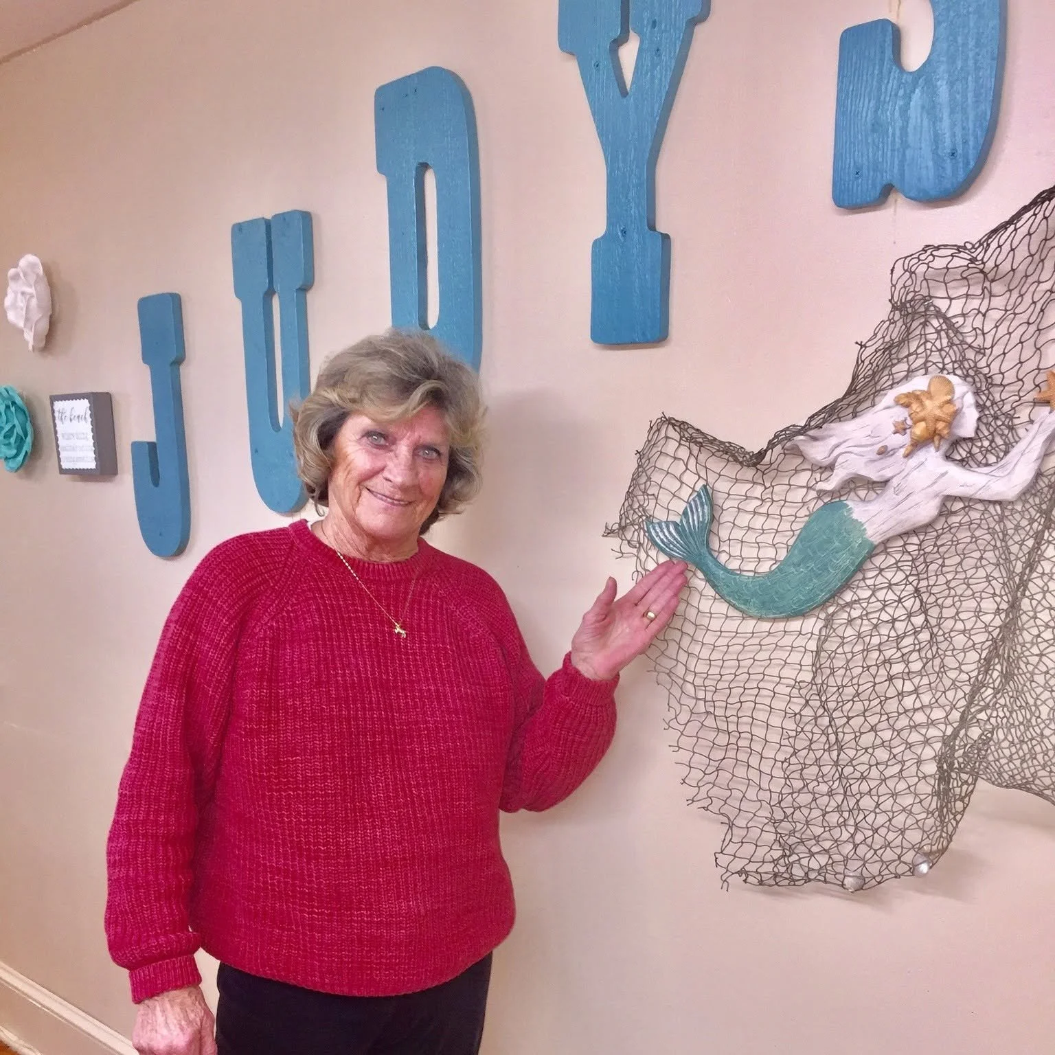 A woman with gray hair wearing a red sweater and black pants is touching a decorative fishing net with a pastel-colored mermaid tail on it. She is smiling and standing in front of a wall with large blue letters.