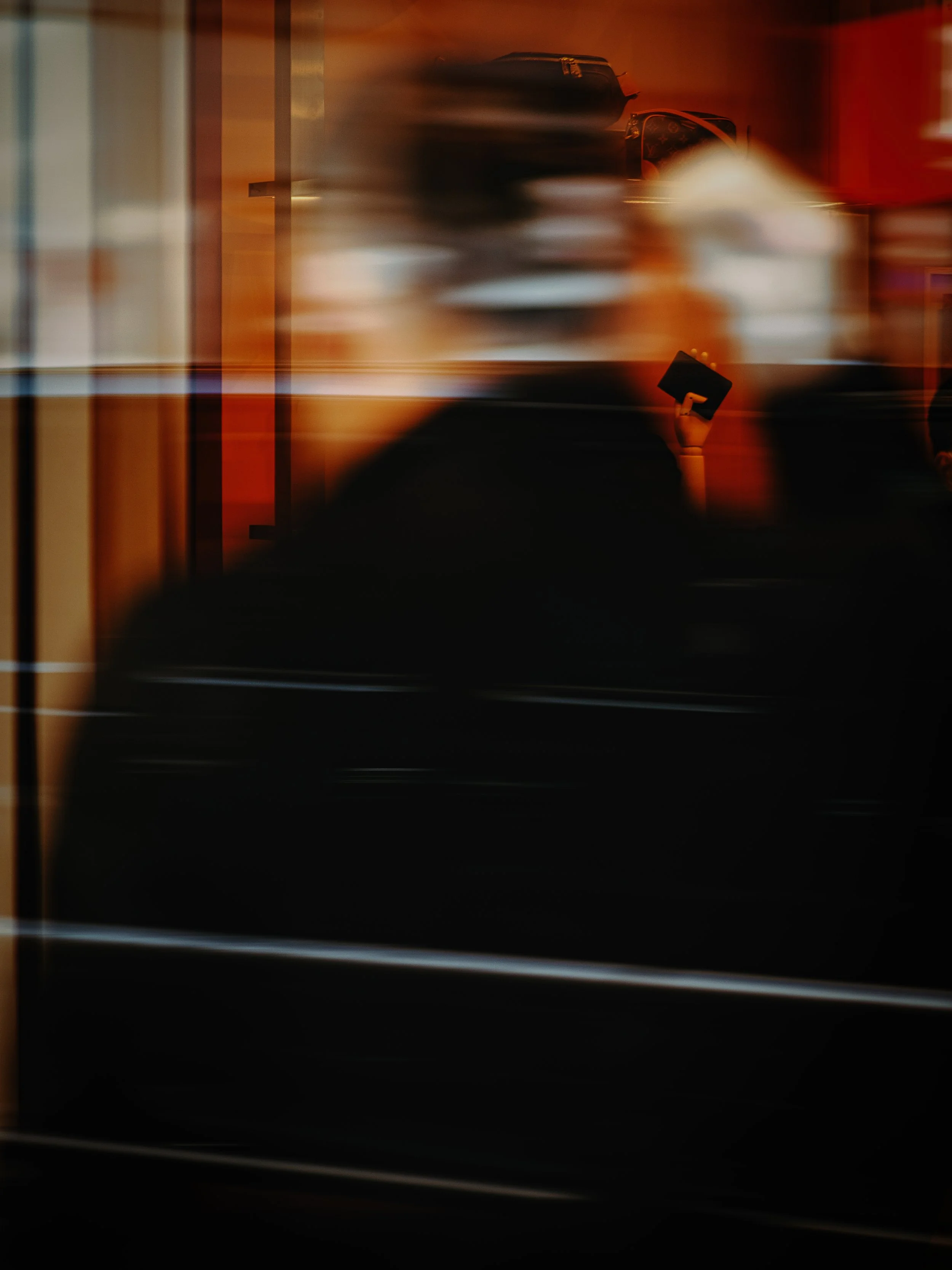 Blurred image with warm orange and red tones showing a hand holding a small black book or notebook, and a background with indistinct objects, possibly in a room or store.