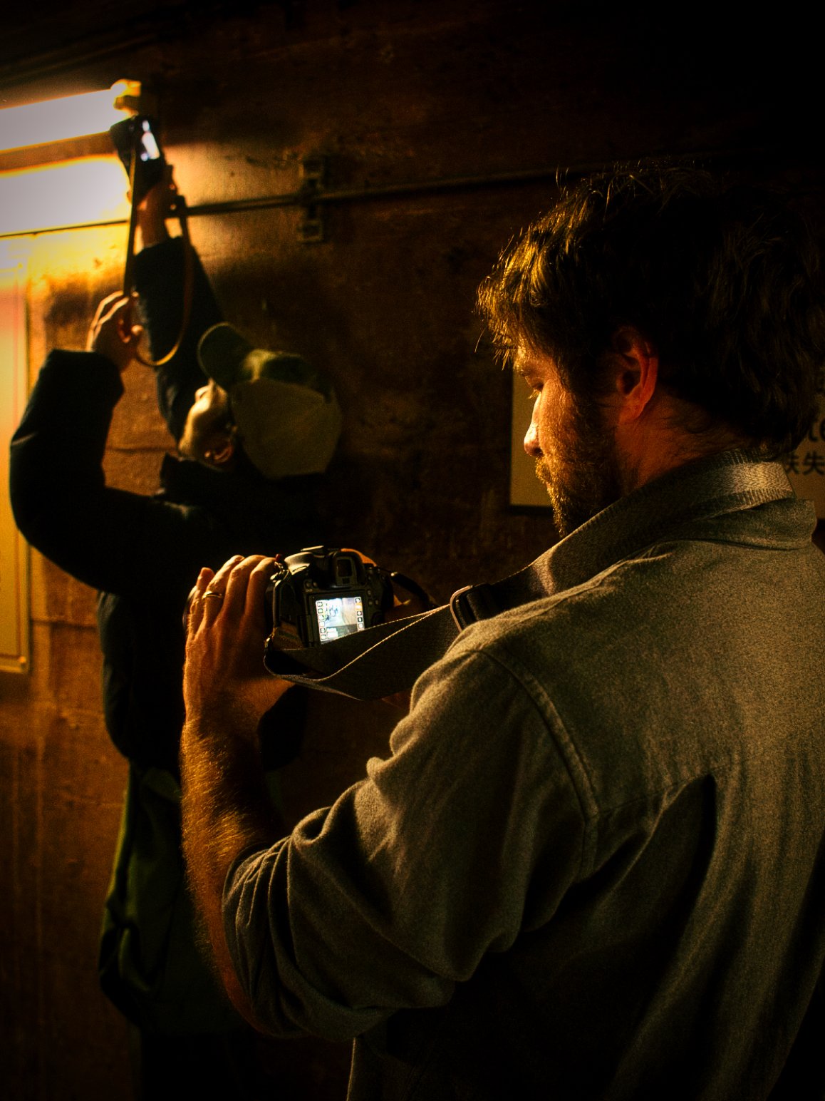 Two men are taking photographs in a dimly lit room, one is holding a camera and looking at its screen, while the other is adjusting his camera on a wall.