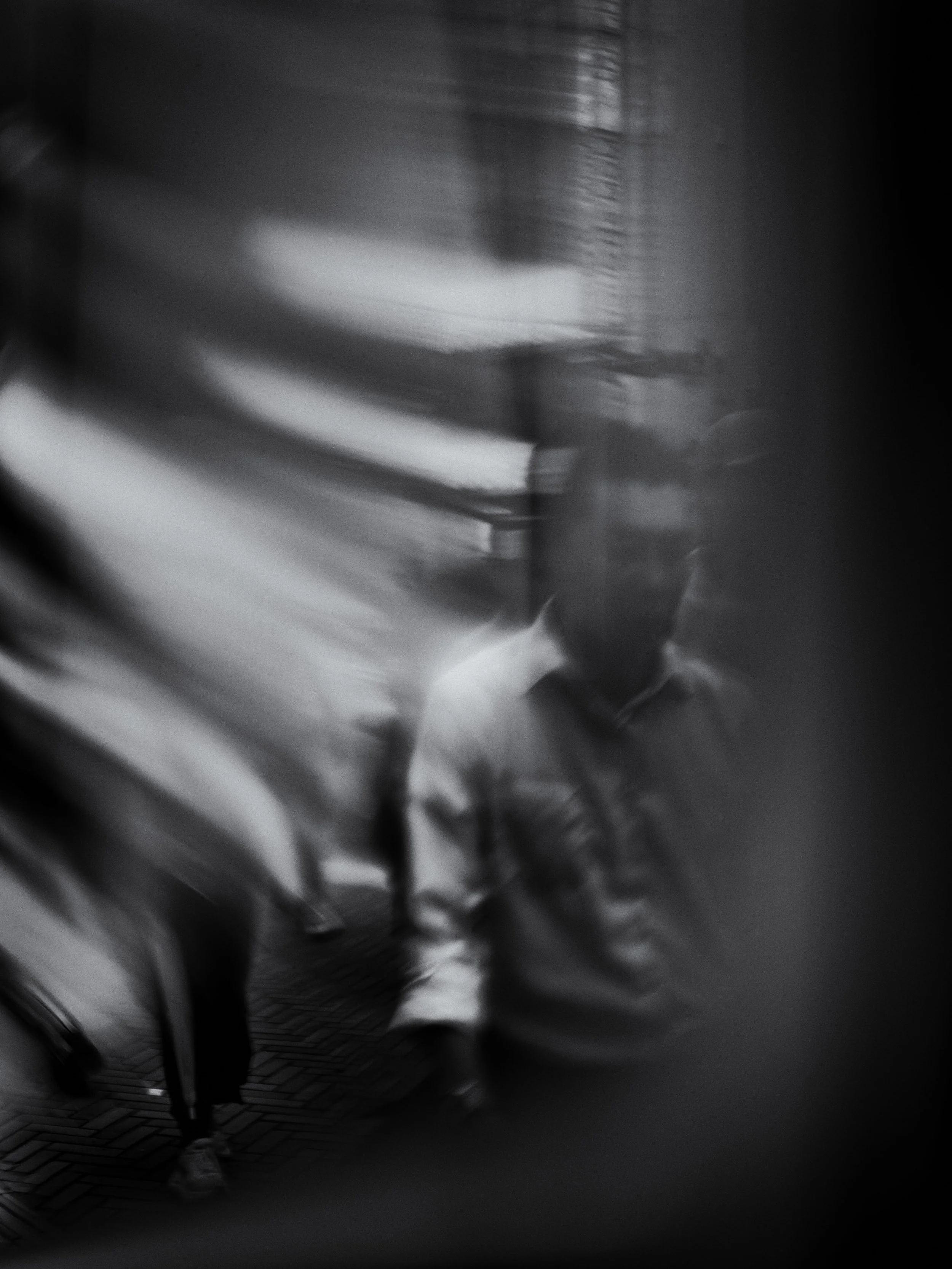 Blurred black and white photo of people walking on a city sidewalk, with buildings in the background.
