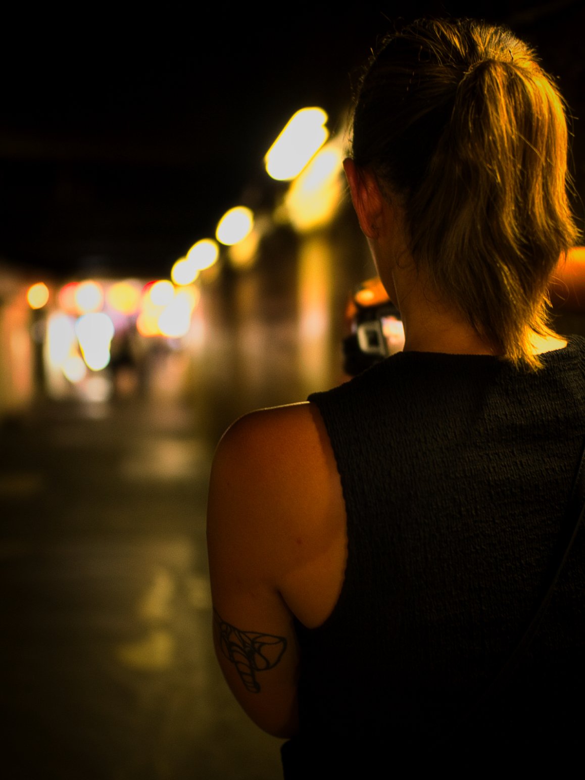 Back view of a woman with blonde hair styled in a ponytail, standing in a dimly lit urban street at night, illuminated by yellow streetlights.