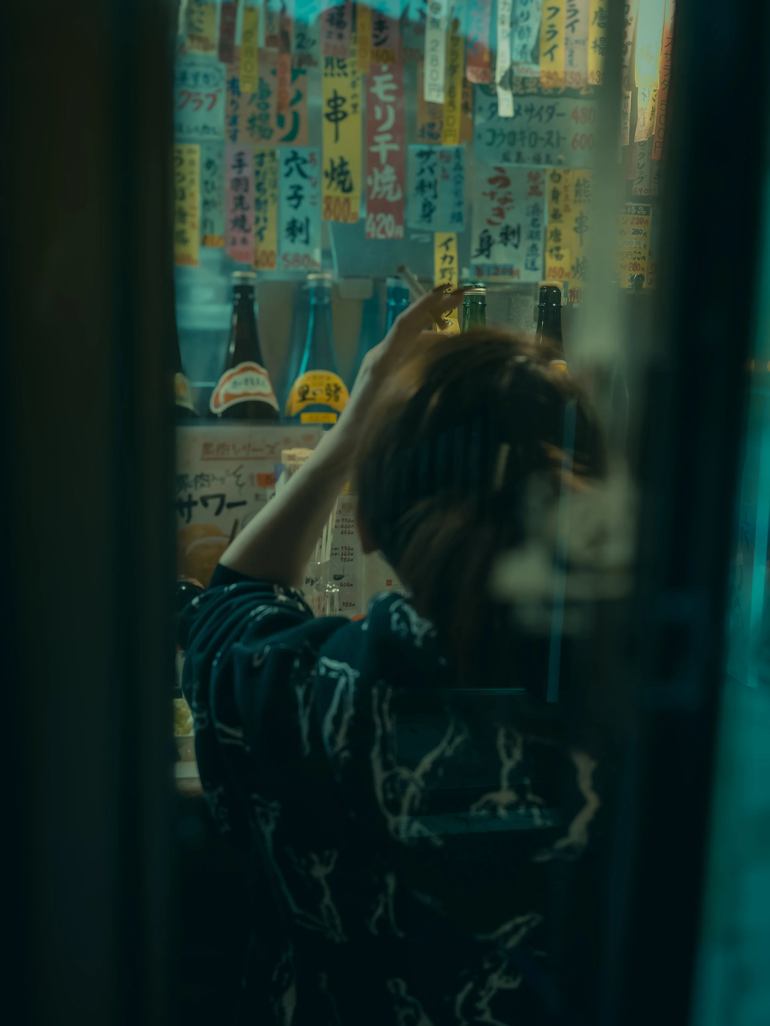 A person with short hair and glasses taking a selfie in front of a mirror covered with colorful Japanese posters and signs, with bottles on the counter.