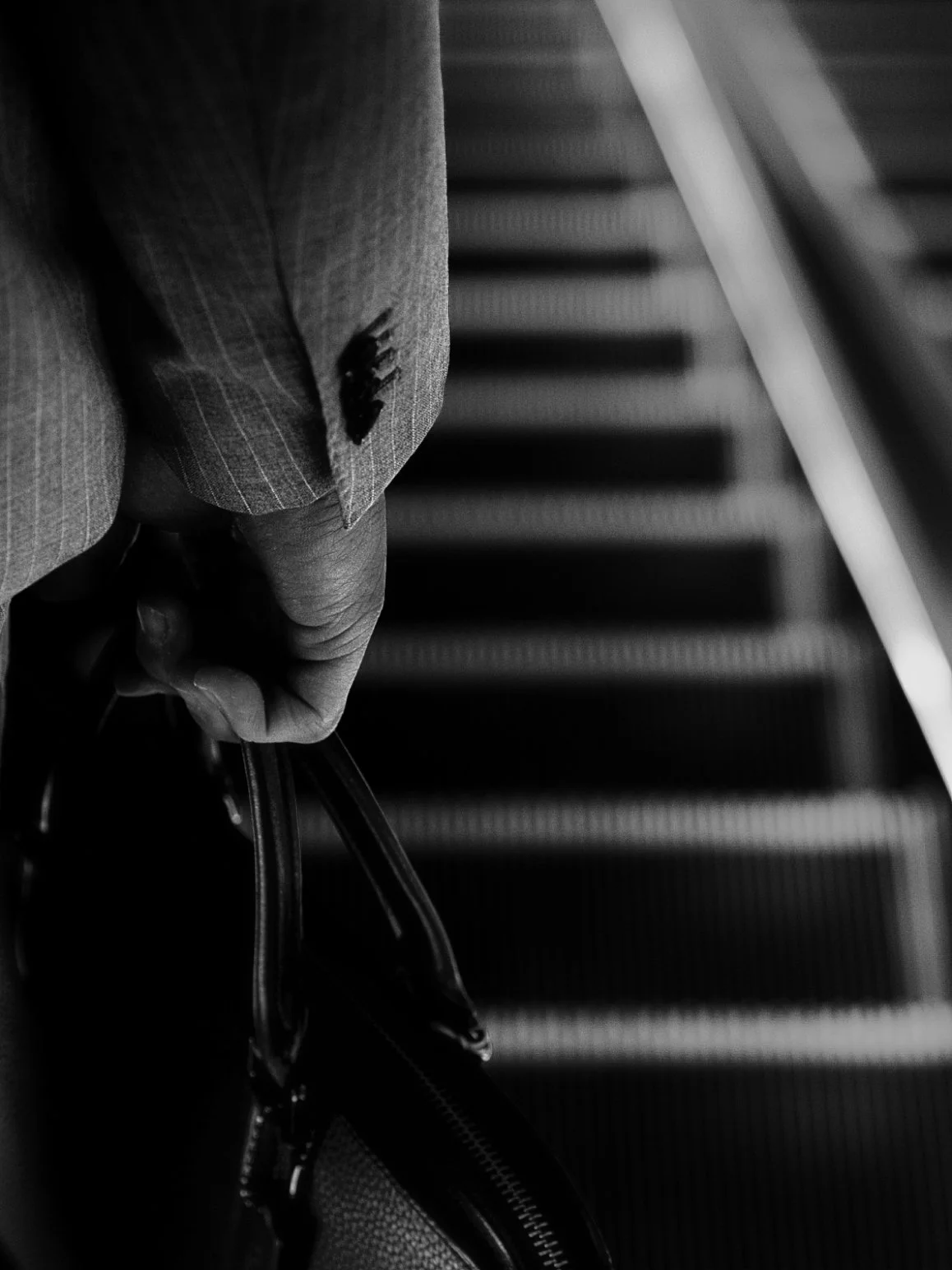 Black and white photo of a person's hand holding a briefcase while walking up an escalator, seen from above.