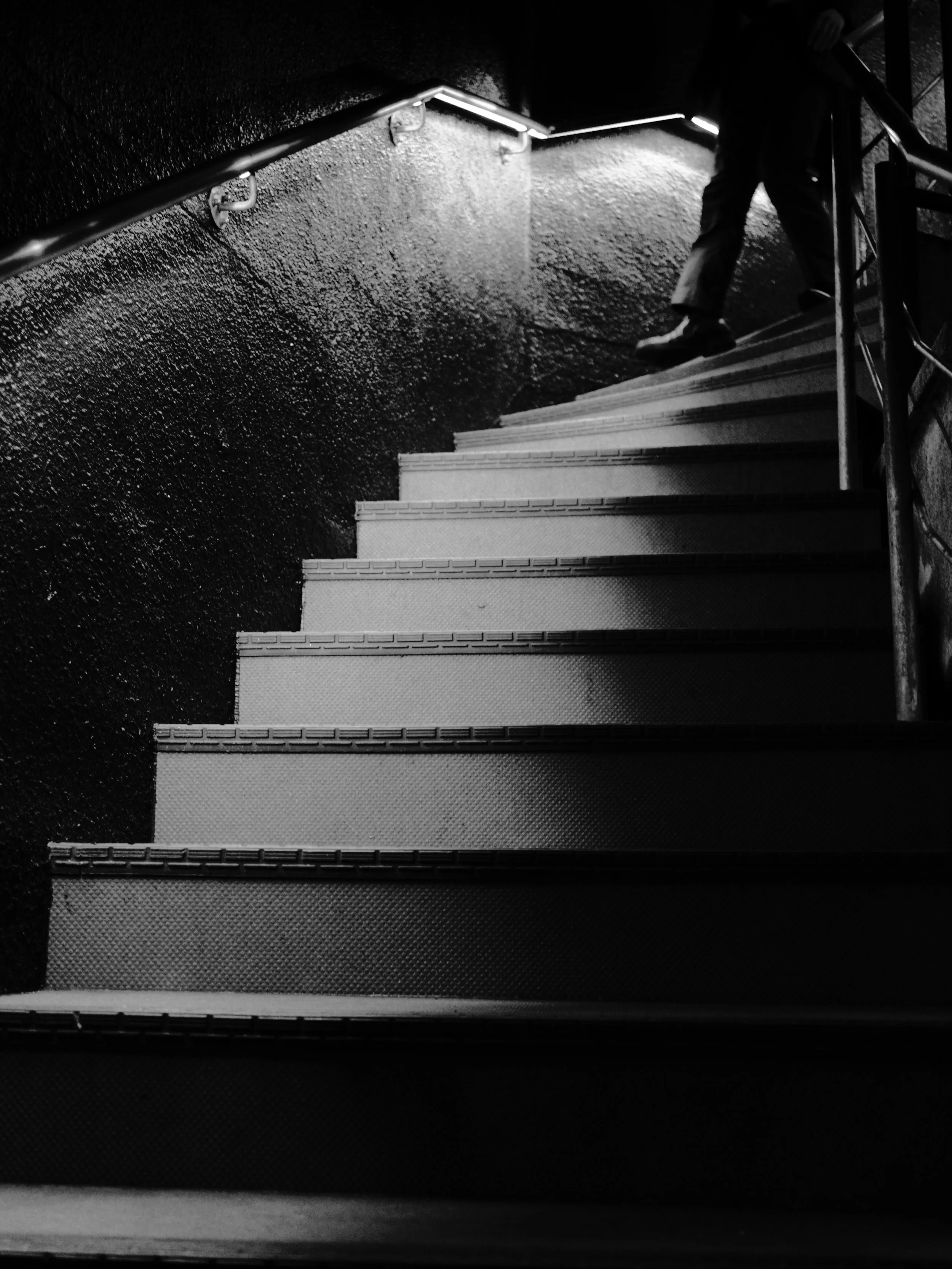 Black and white photo of a person walking down a staircase at night, taken from the bottom of the stairs showing only their legs and feet.
