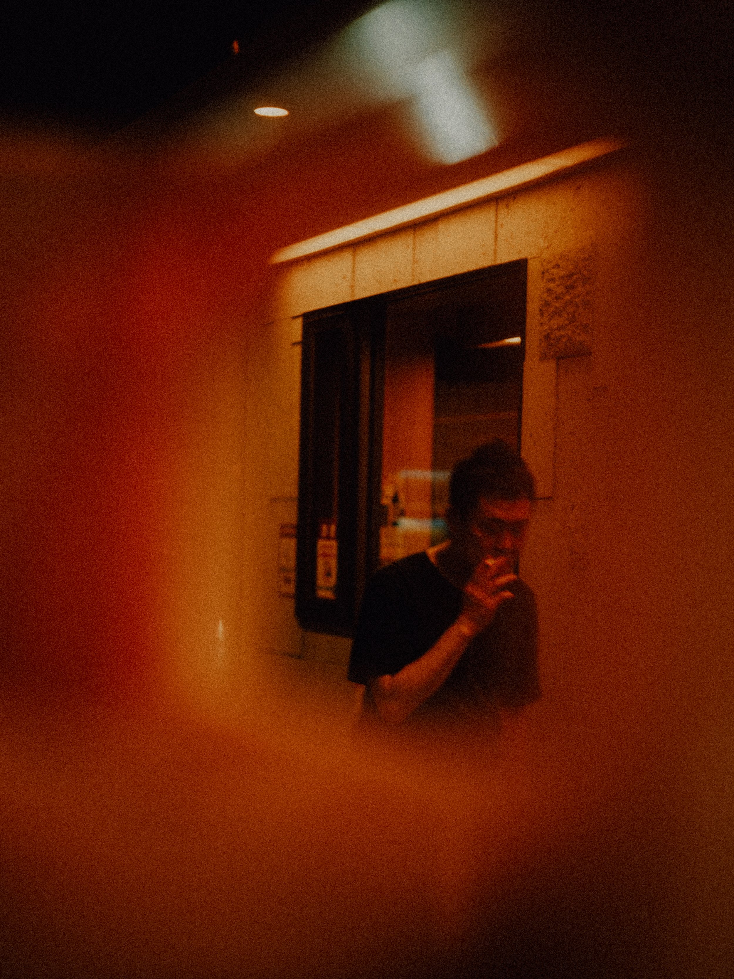 A person with short hair, glasses, and a dark shirt is taking a selfie in a dimly lit room, holding a drink in front of their mouth and smiling.