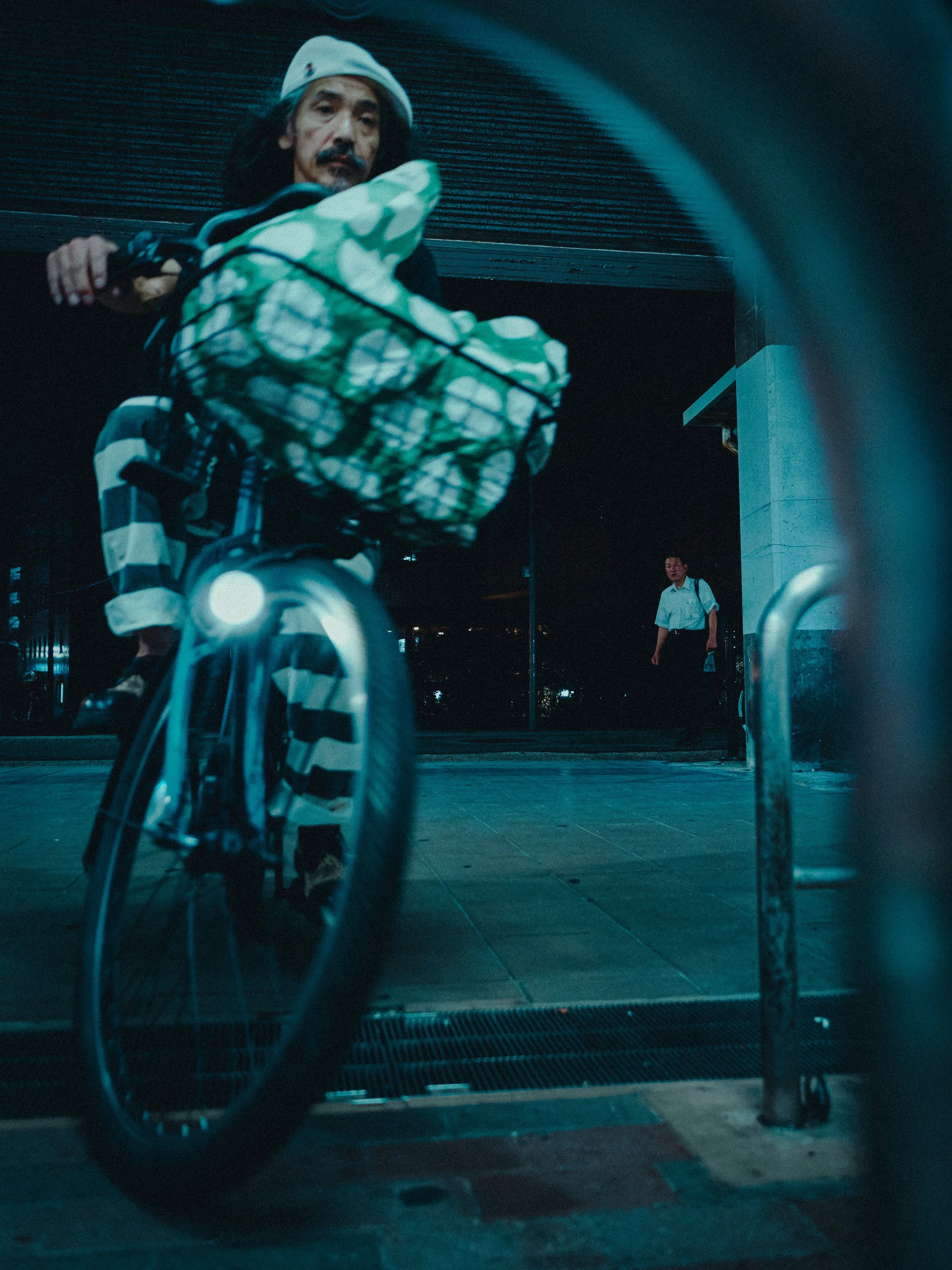 View of a person with long hair, a mustache, and a white cap, riding a bicycle with a camouflage-patterned bag, seen through a low-angle, slightly blurry shot taken from behind a glass door or window, with a man in a white shirt and backpack in the b