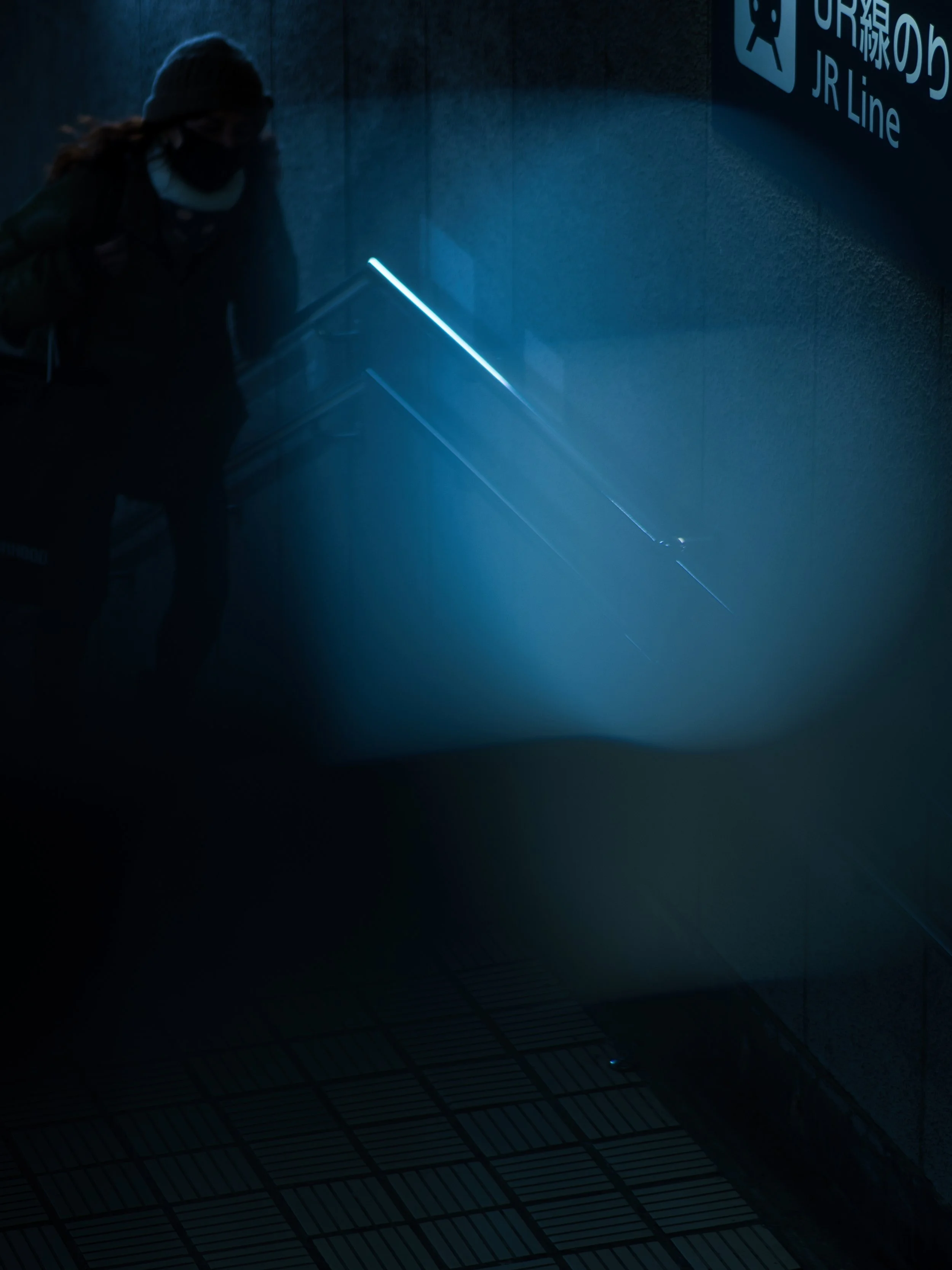 A person wearing a face mask, beanie, and jacket walking in a dimly lit indoor space near a sign for JR Line, with blue lighting and a tiled floor.