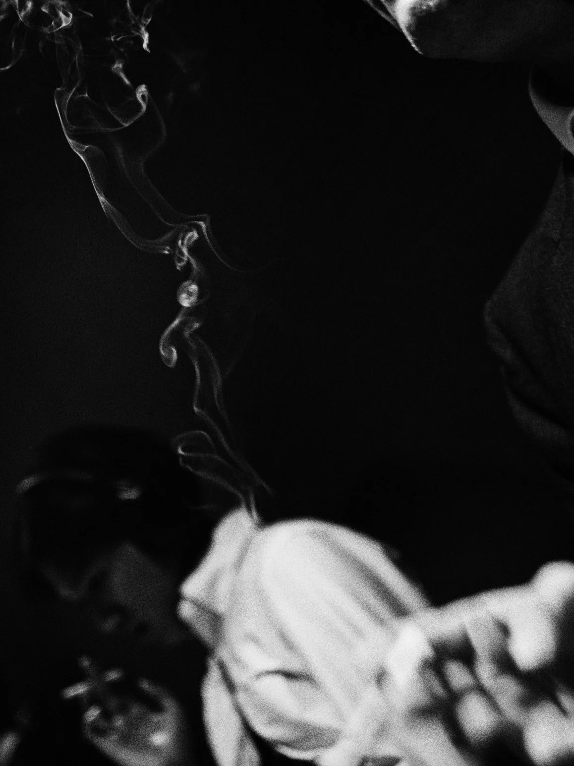 A close-up black and white photo of the tips of two cigarettes with smoke rising.