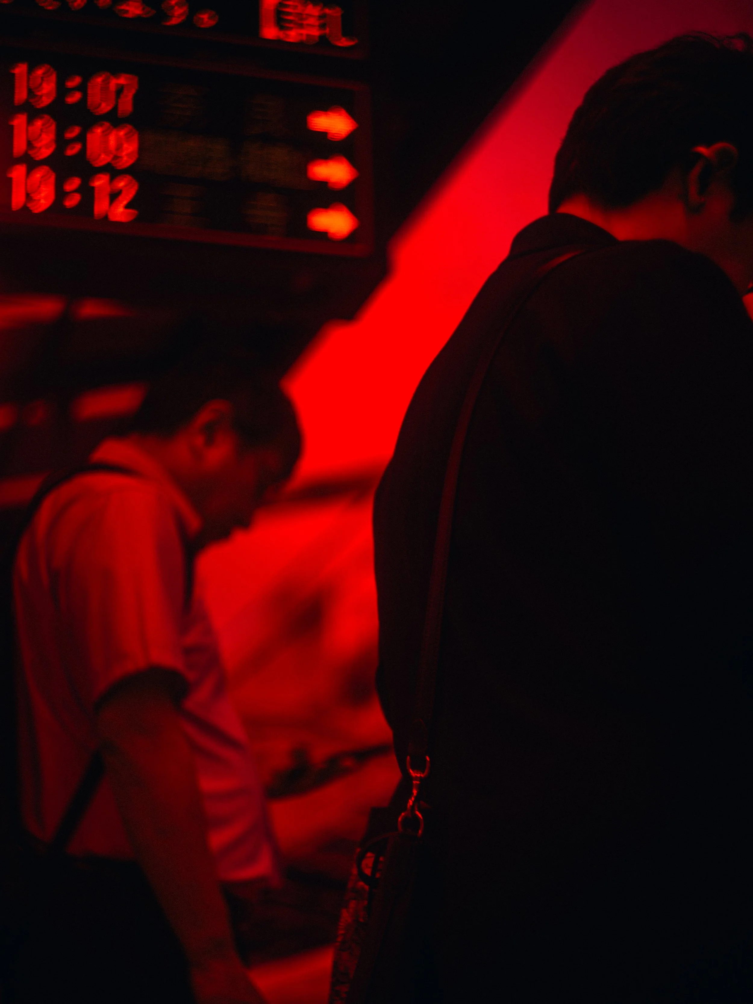 A person wearing glasses looking down while inside a room illuminated with red light, with a digital timetable or display showing times and arrows on the wall in the background.