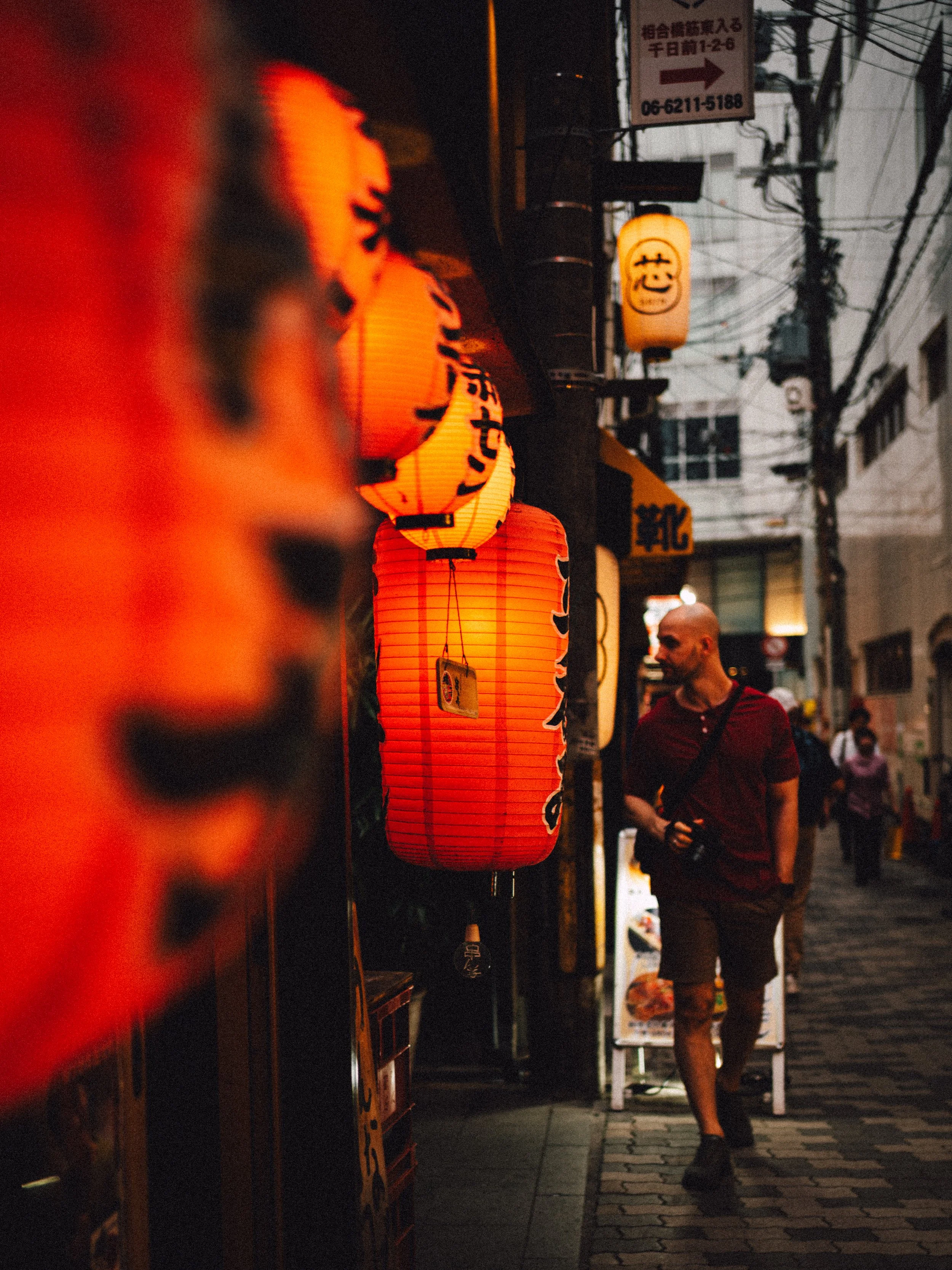 A street scene at dusk with Japanese paper lanterns hanging along the sidewalk, and a man in a red shirt and shorts walking past. There are electrical wires overhead and signs in Japanese on the buildings.