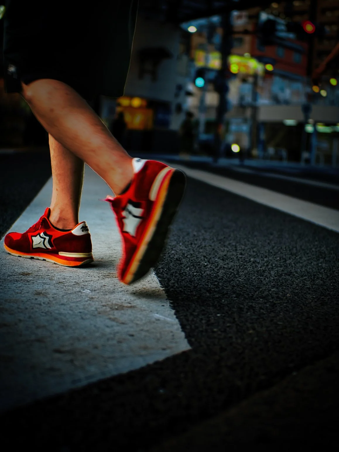 Close-up of a person wearing red sneakers crossing a street at night, with city lights and buildings blurred in the background.