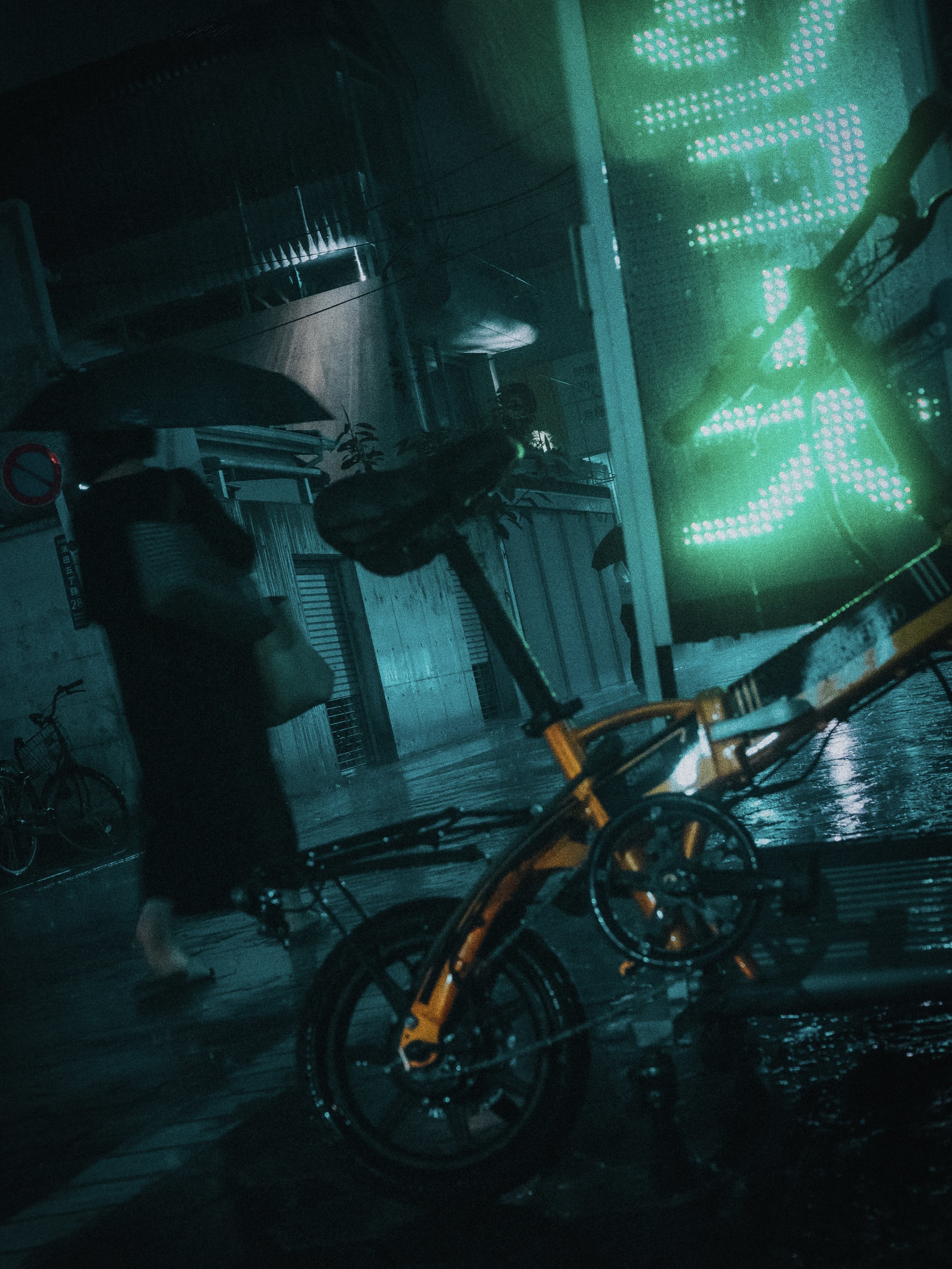 Nighttime scene showing bicycles, umbrellas, and a person walking with an umbrella, reflecting wet pavement, illuminated by a large green digital display.