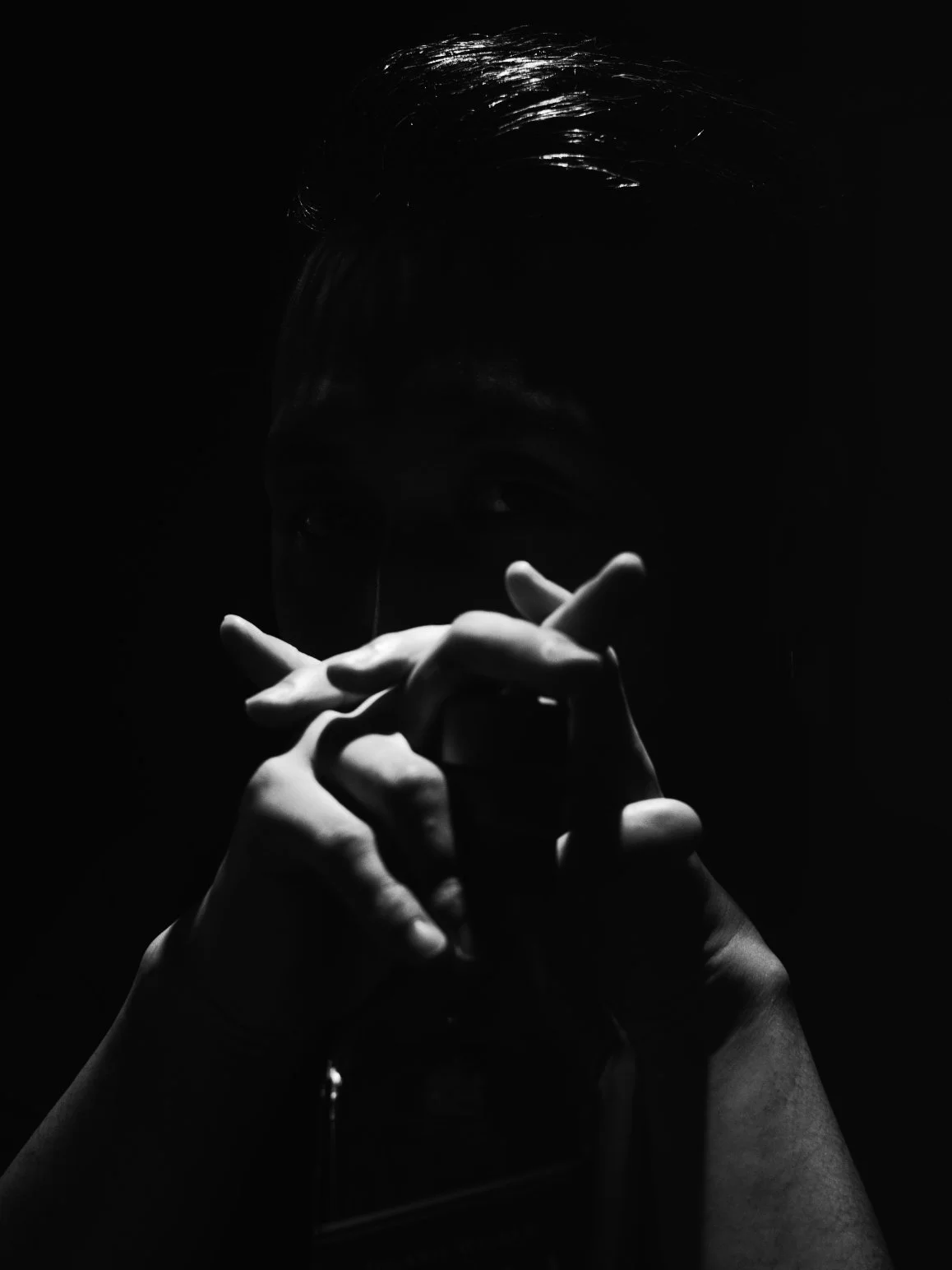 A person with slicked-back hair, partially illuminated, is pointing fingers towards the camera in a dramatic pose against a dark background.