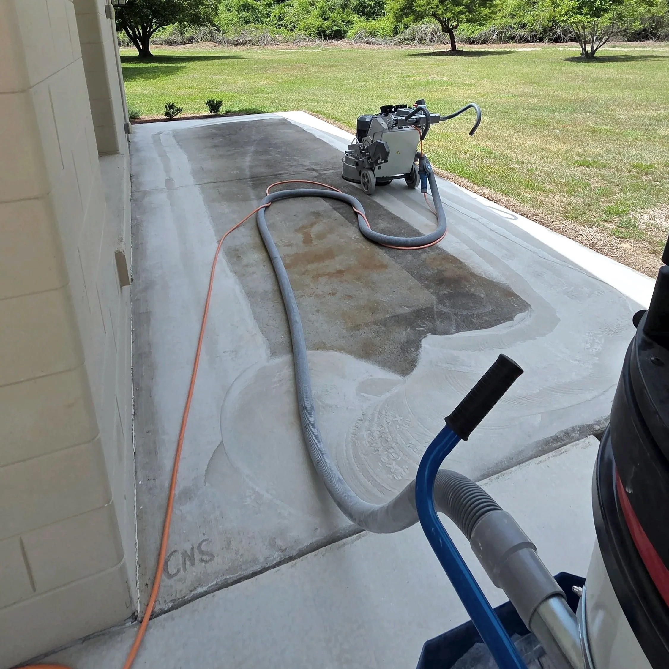 A concrete patio with a pressure washer placed on it, connected with hoses. The patio is next to a grassy yard with trees in the background.