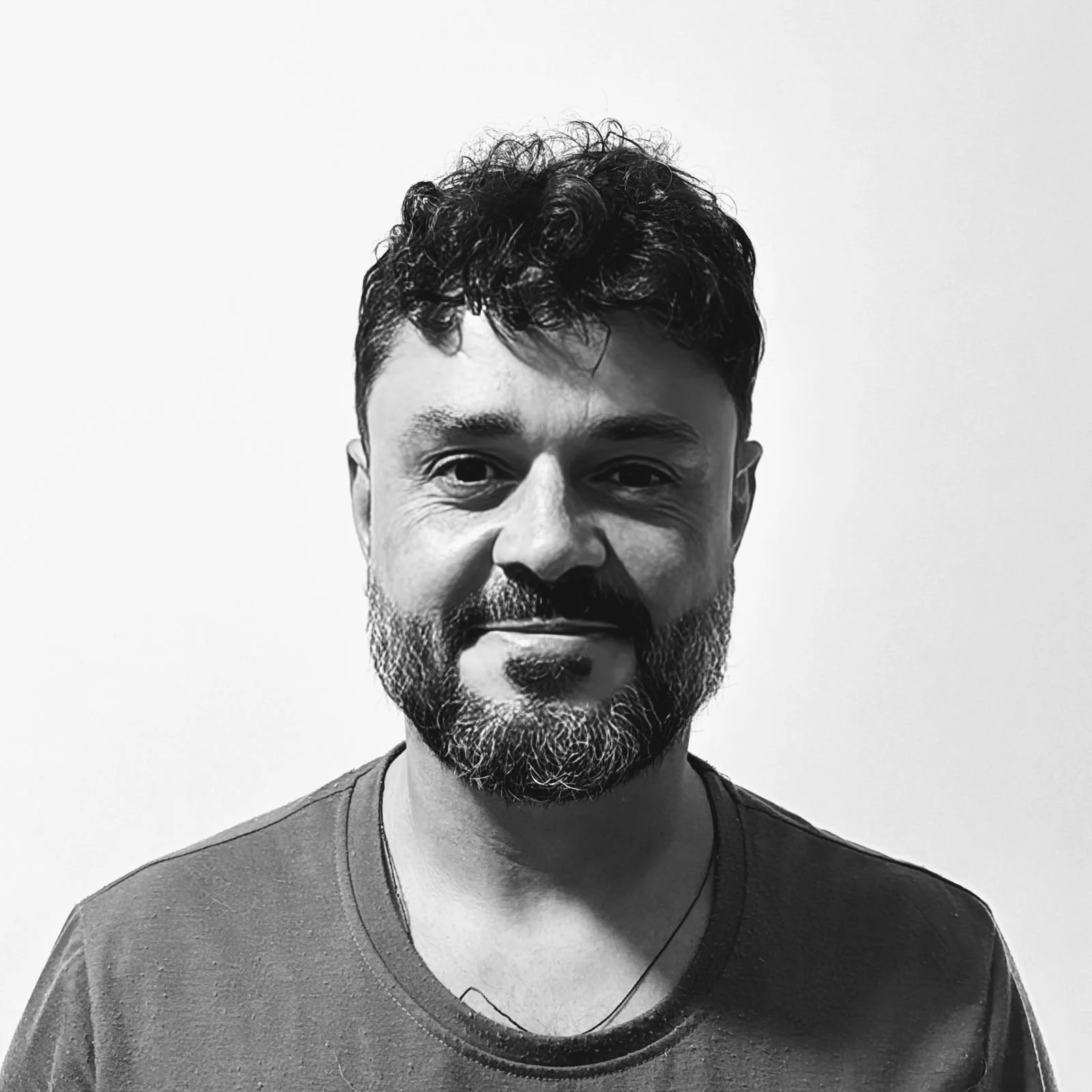 A black-and-white portrait of a man with curly hair and a beard, smiling slightly, wearing a t-shirt and earphones.