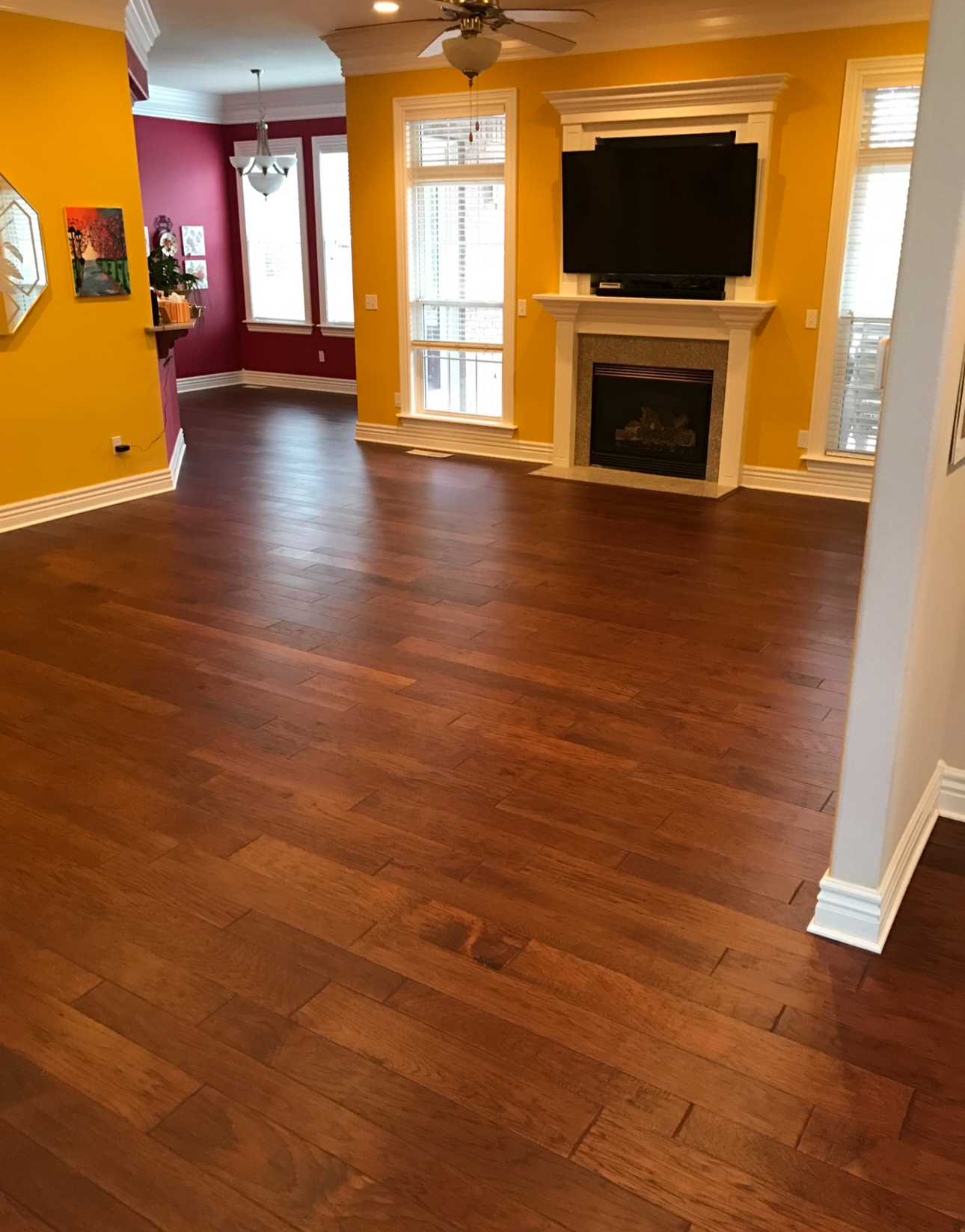 Living room with dark hardwood floors, yellow walls, a fireplace with a mounted TV above it, and windows with white blinds. Part of a dining area with red walls and a chandelier is visible in the background.