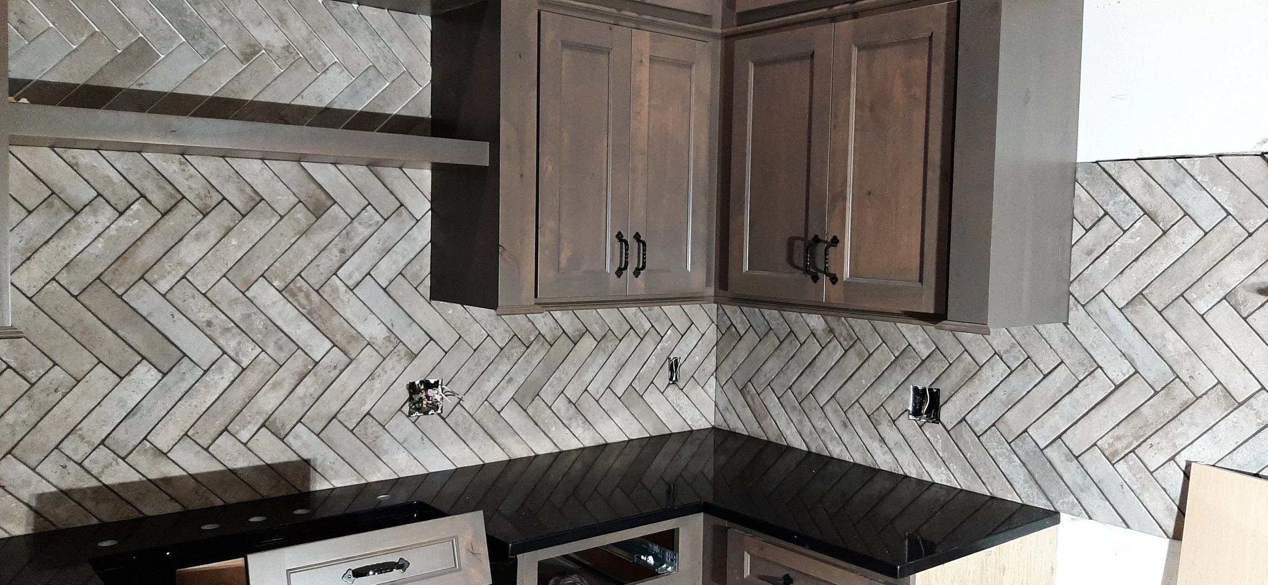 Kitchen with herringbone tile backsplash, dark wood cabinets, black countertop, and electrical outlets