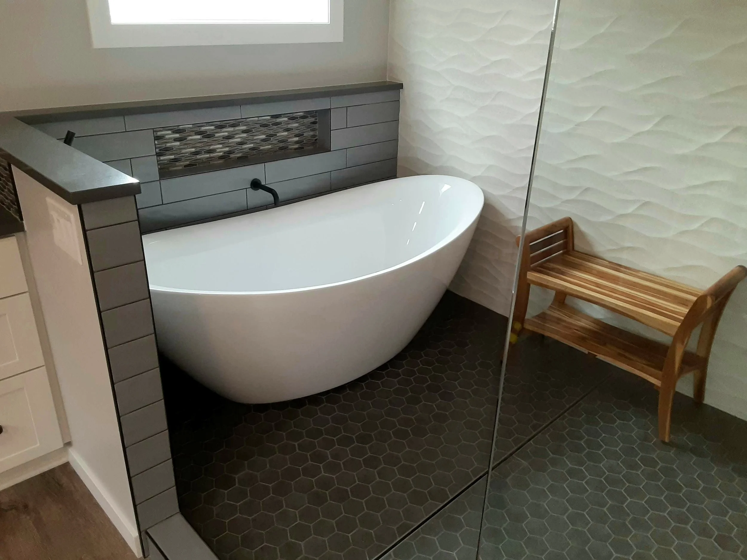 A bathroom with a white freestanding bathtub positioned against a tiled wall with a shelf and mosaic accent. To the right, there are wooden chairs and a glass partition. The floor is covered in dark hexagonal tiles, and a window is visible above the bathtub.